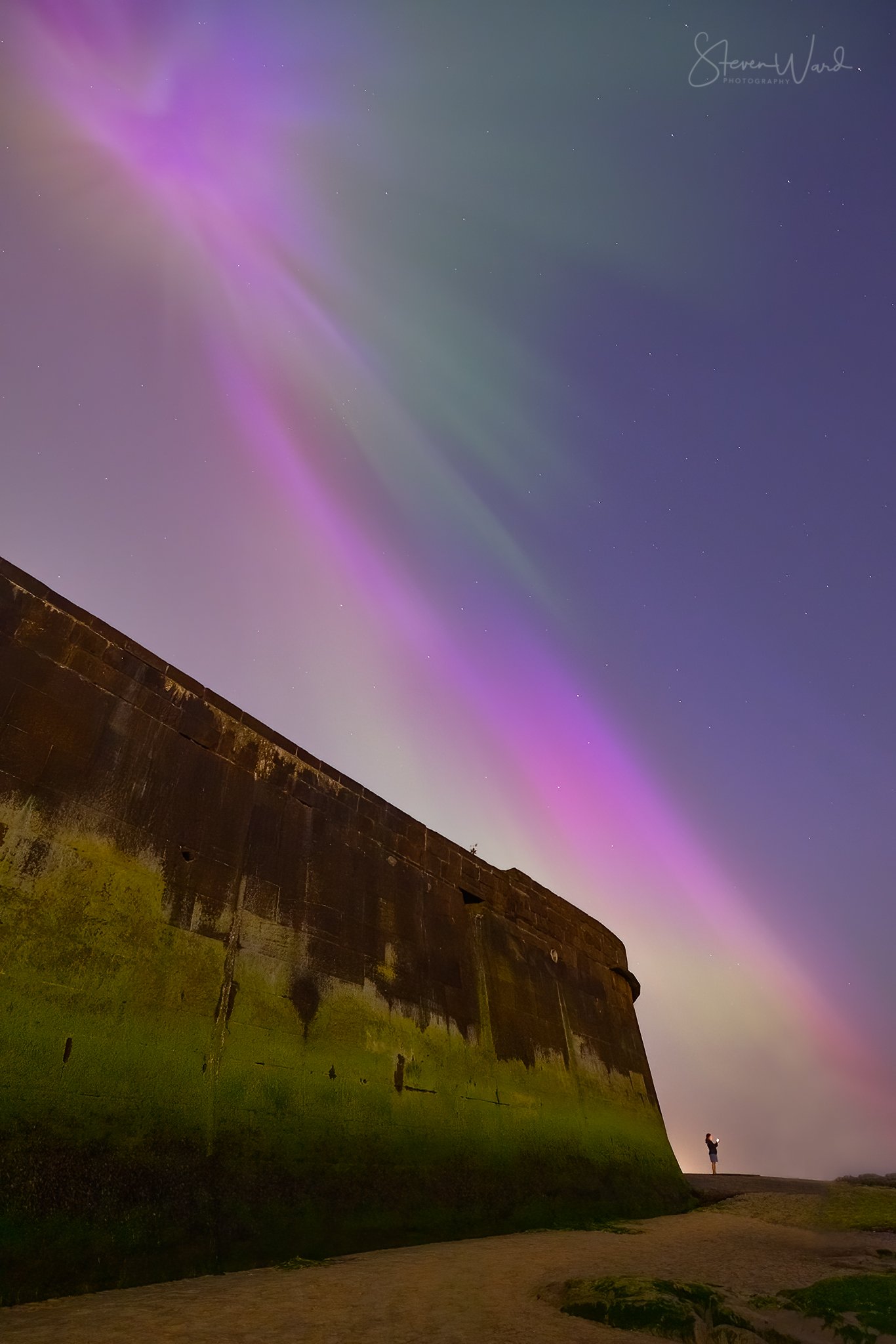 Night sky with colorful aurora and stars, with a large weathered concrete fortress wall in the foreground and a person standing near the base of the wall taking a photo.