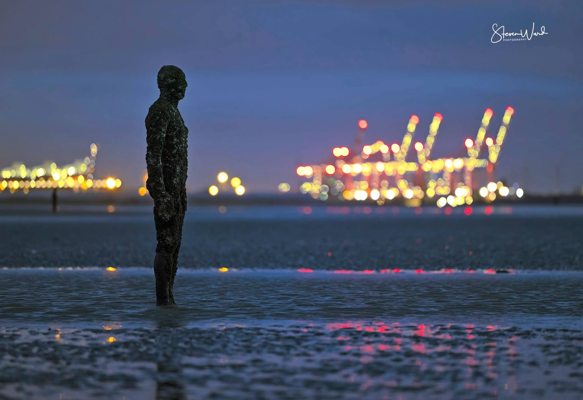 Statue of a man standing in water at night with a port and cranes illuminated in the background.