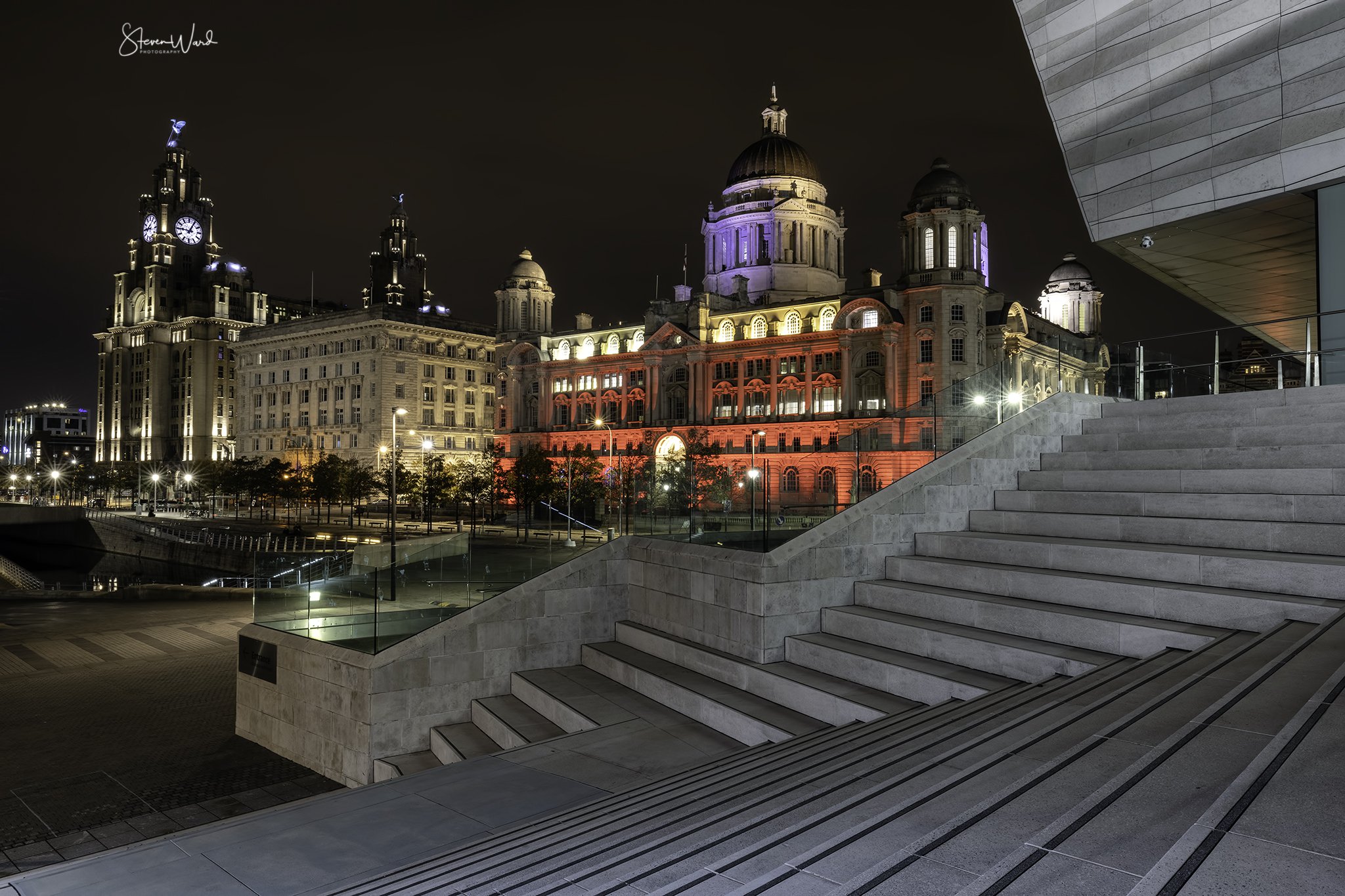 Night view of a historic dome building illuminated with red and purple lighting, with tall clock towers on either side, seen from a modern staircase with glass railing in the foreground.