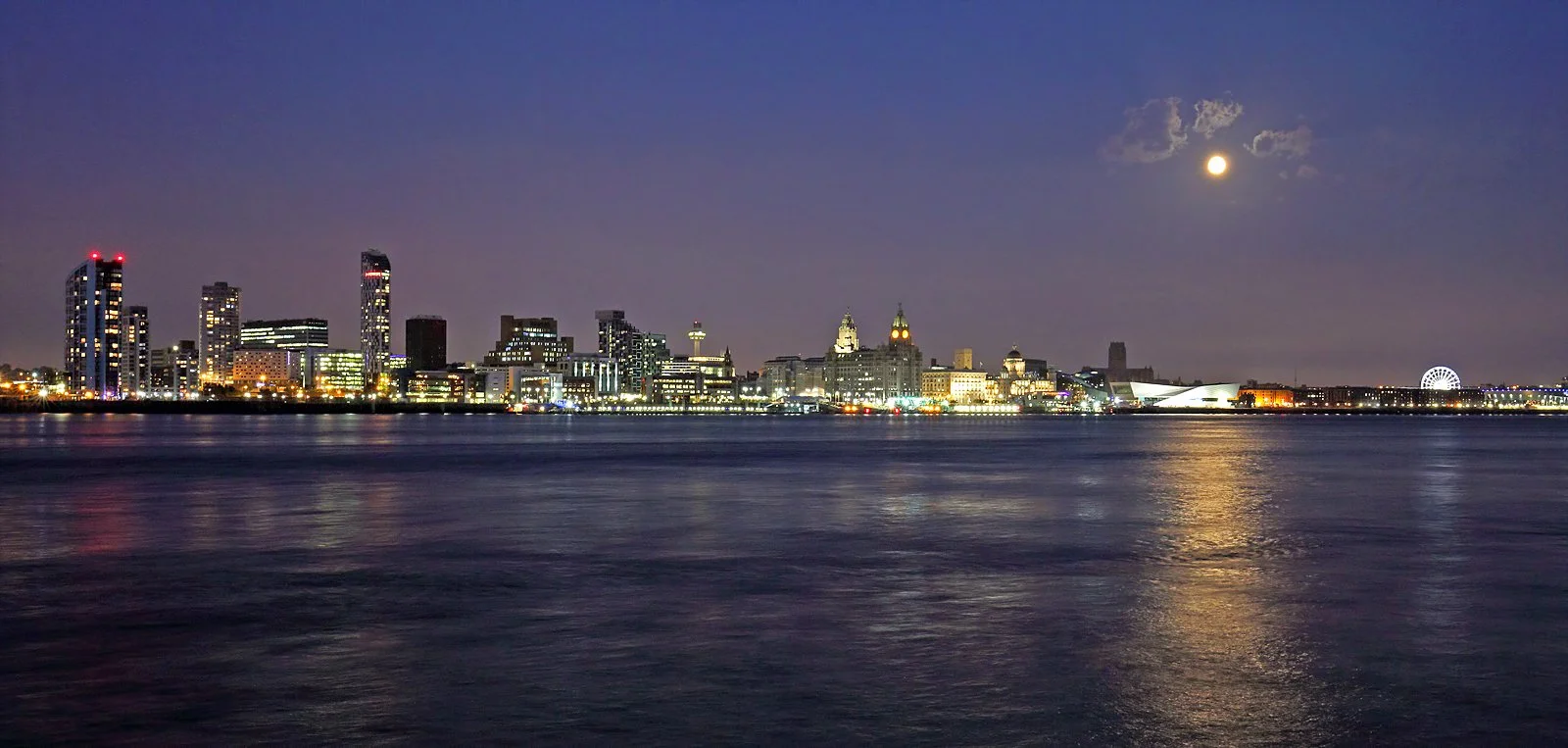Nighttime city skyline with illuminated buildings, reflecting on calm water, under a bright full moon with some clouds.