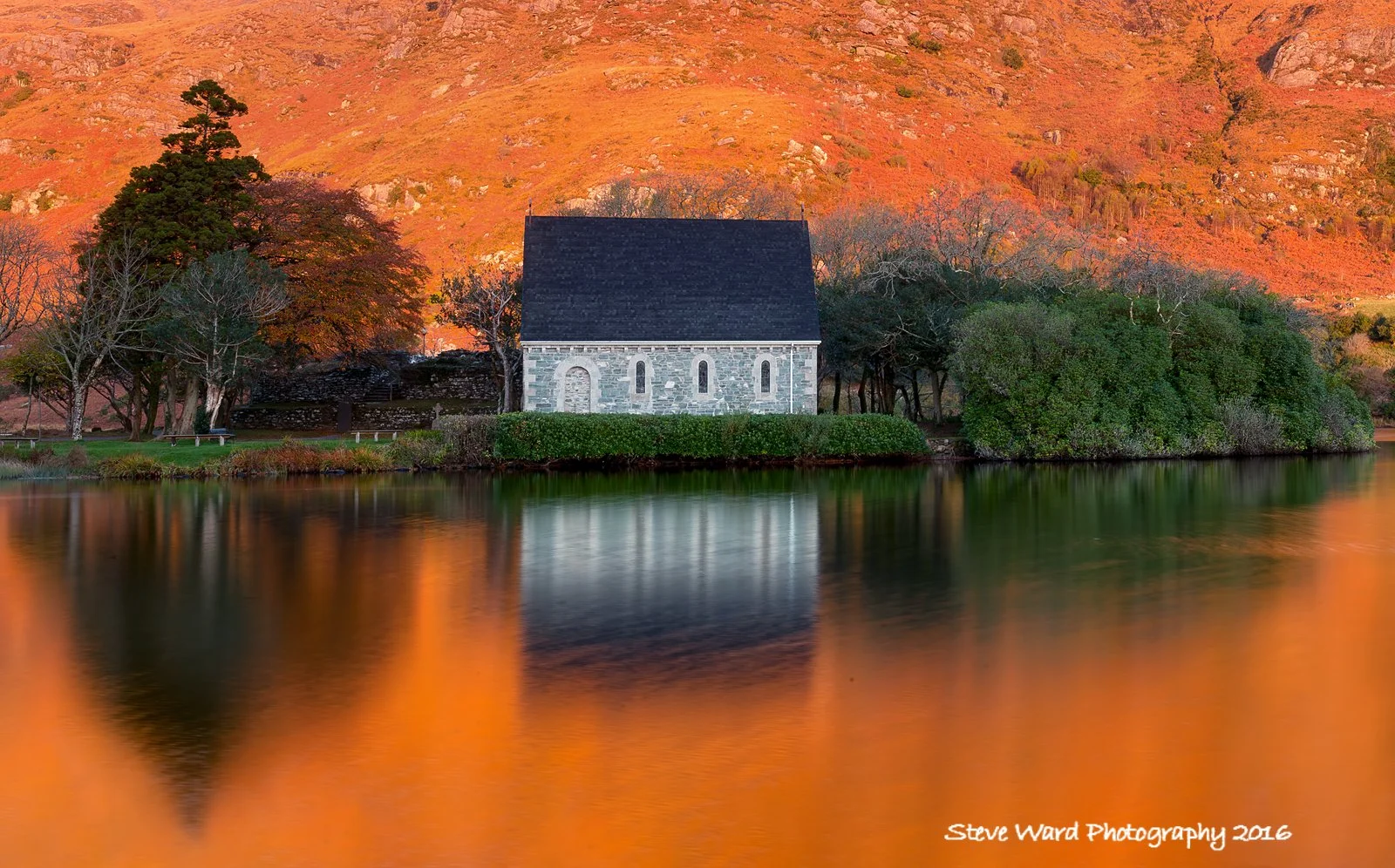 A small stone church with a black roof reflected in a lake, surrounded by trees with orange foliage, with a mountain in the background during sunset.
