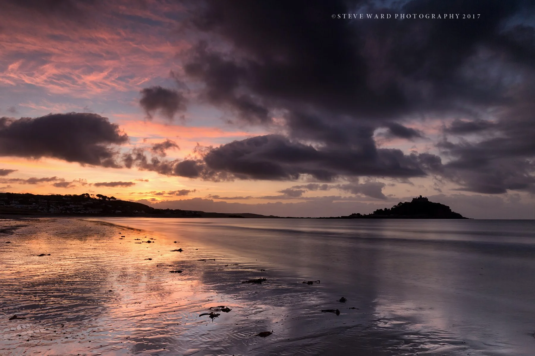Sunset over a calm beach with clouds in the sky and a small island with a building on it in the distance.