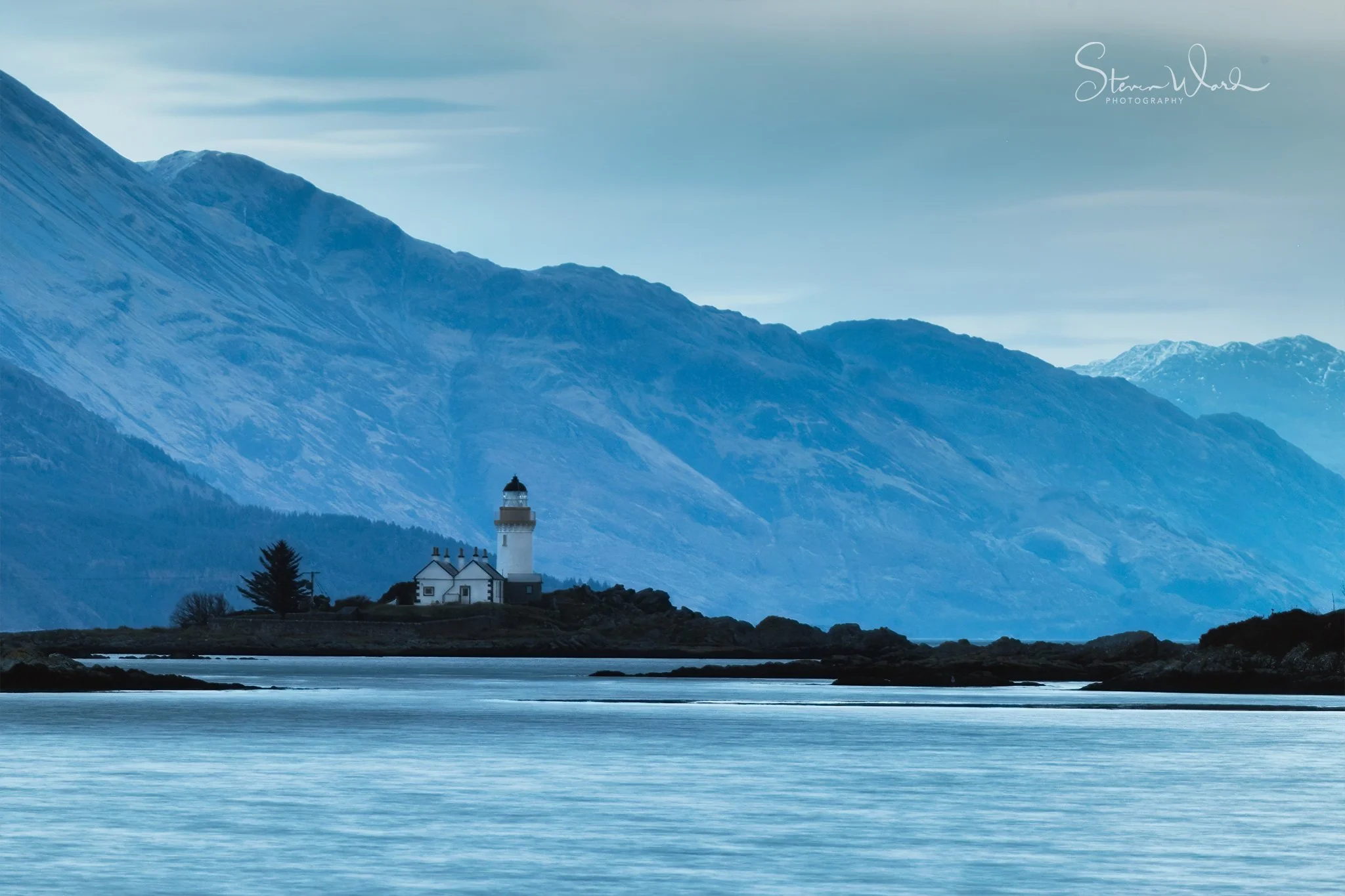Lighthouse on a small rocky island near calm water with mountains in the background, under a cloudy sky.