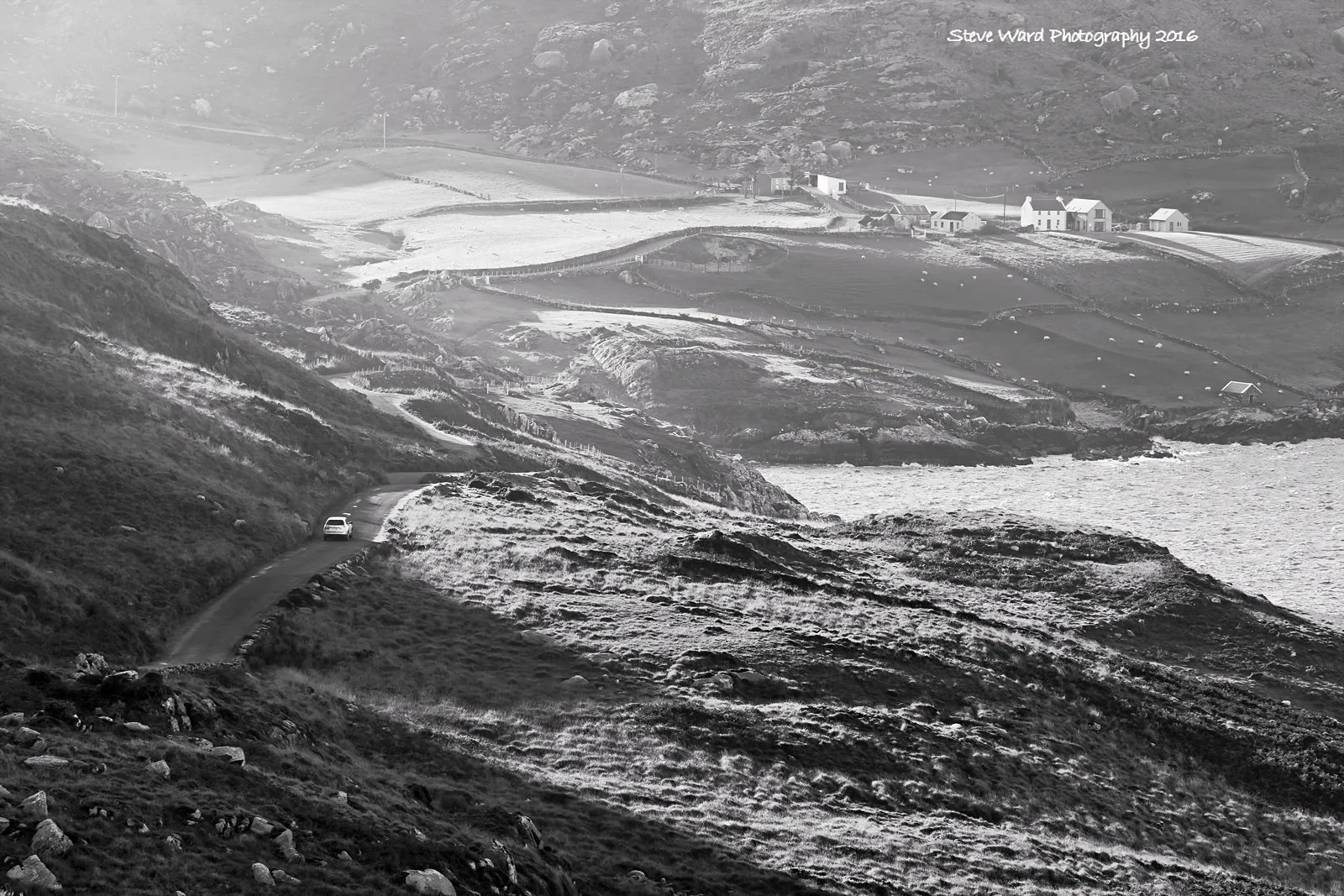 Scenic coastal landscape in black and white featuring rolling hills, a winding road with a car, and a cluster of houses in the distance.
