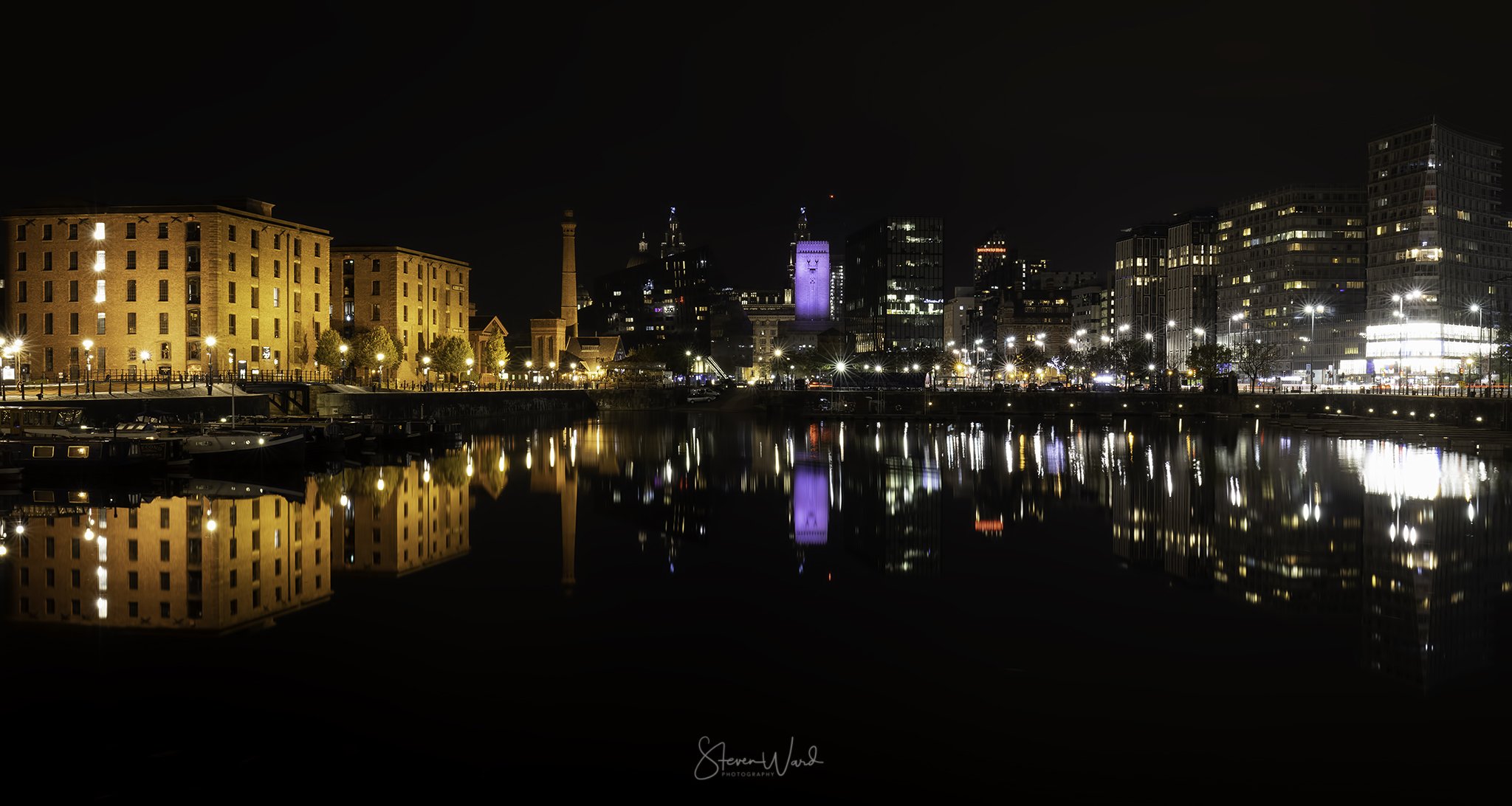 Nighttime city skyline reflected in water with illuminated buildings and streetlights.