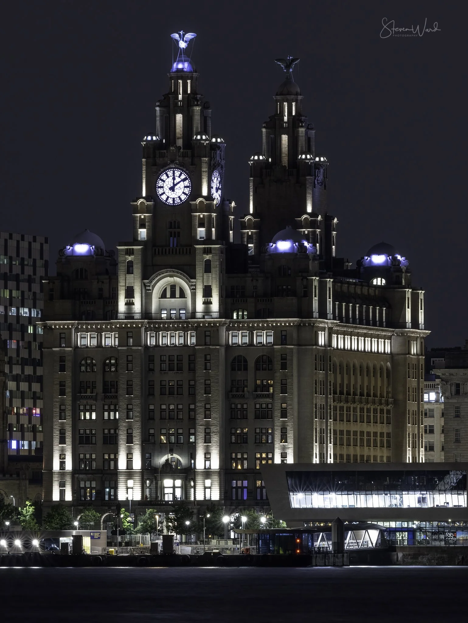 Night view of a historic building with two clock towers and illuminated statues on top, with surrounding modern structures and a glass-covered walkway in the foreground.