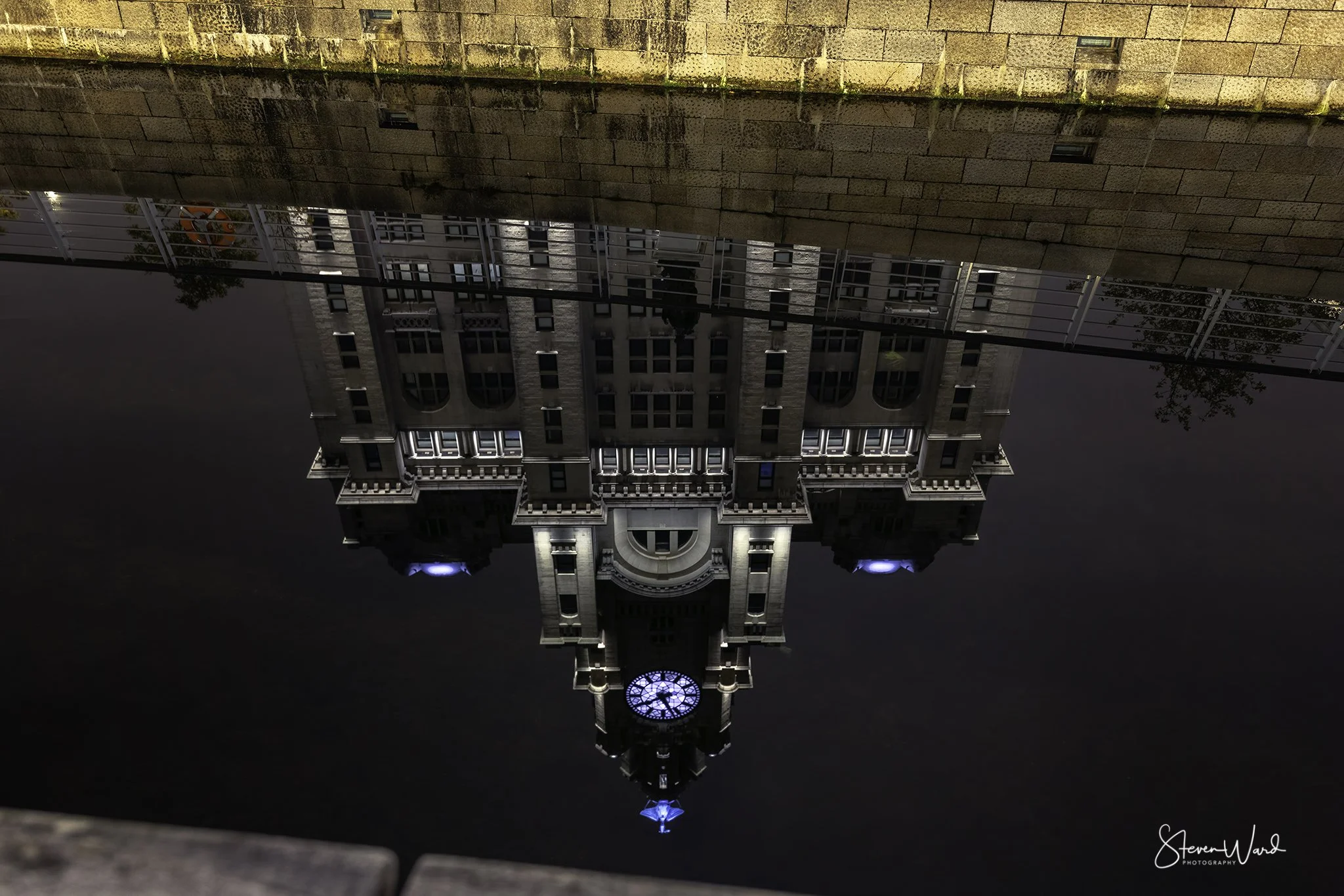Upside-down view of an ornate building with a clock tower at night, reflected in a dark body of water.
