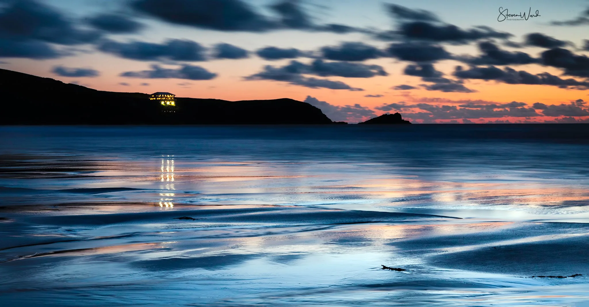 A scenic view of a beach at sunset with calm waves, a silhouetted headland, and a colorful sky with clouds.