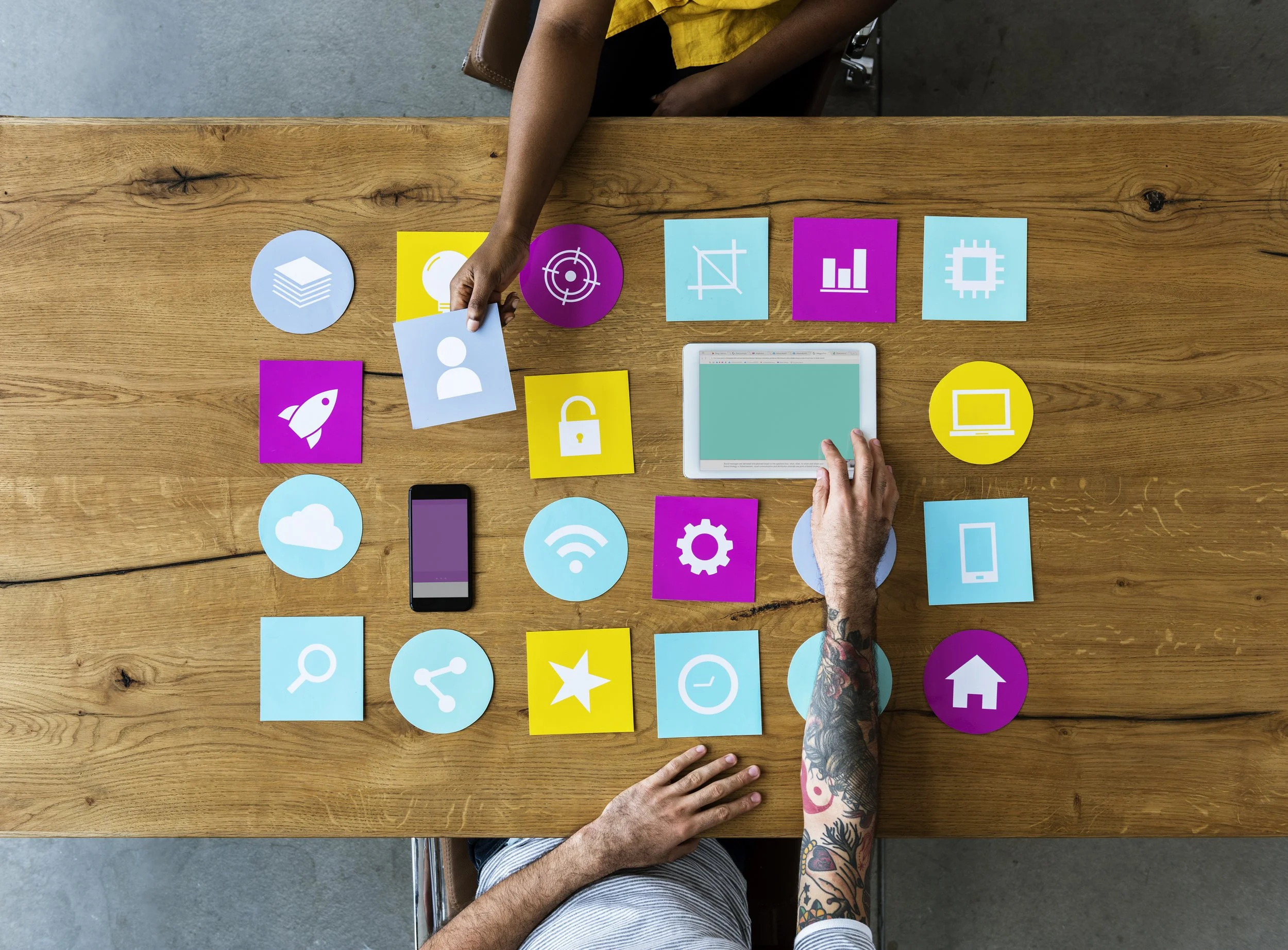 Top-down view of two people arranging colorful digital icons on a wooden table, including symbols for cloud storage, Wi-Fi, search, settings, home, and a tablet and smartphone.