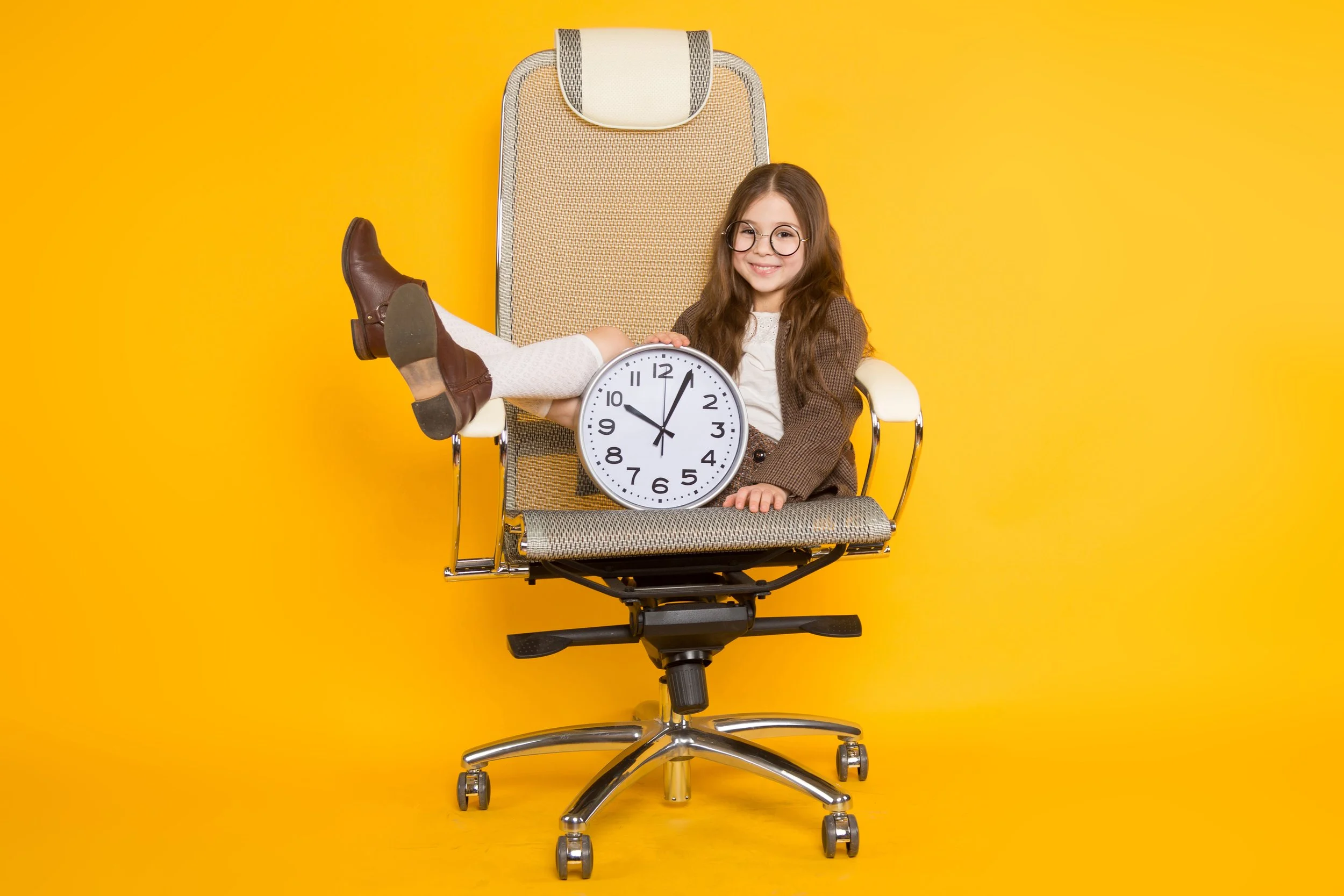 A young girl with glasses sitting on an office chair, holding a large clock in her lap, with her legs resting on the armrest. She is smiling and wearing a brown jacket, white tights, and brown boots. The background is solid yellow.