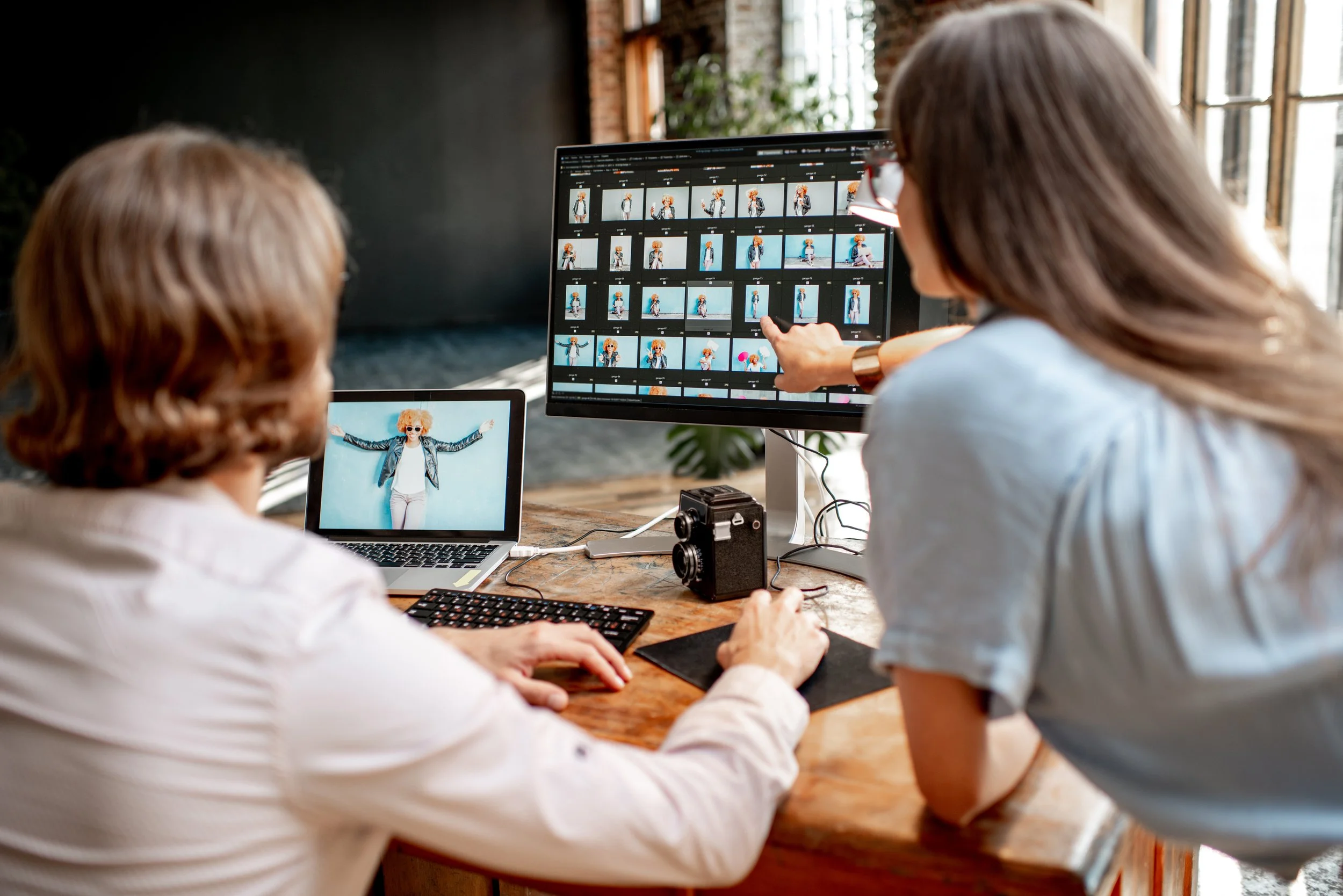 Two women working on photo editing at a wooden desk with computer monitors, a camera, and a laptop, reviewing a photoshoot of a woman posing with sunglasses and a leather jacket.