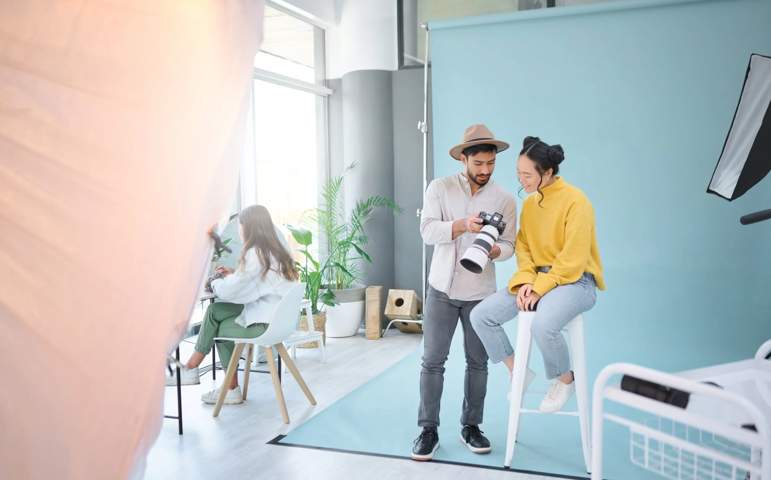 Two people, a man in a light shirt and a woman in a yellow sweater, are in a photography studio. The woman is sitting on a stool with her right leg crossed over her left, and the man is showing her something on his camera. There is a large light shield in the foreground and a blue backdrop behind them. Another person is sitting at a desk in the background.