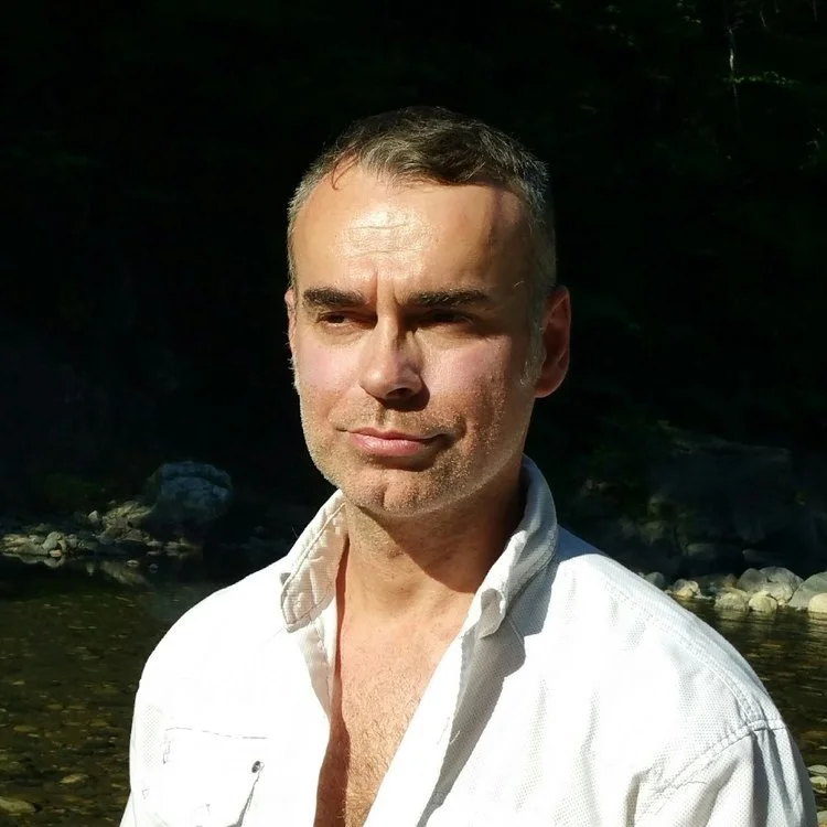 A man with short hair and a slight beard, wearing a white shirt, standing outdoors near a body of water with rocks and trees in the background.