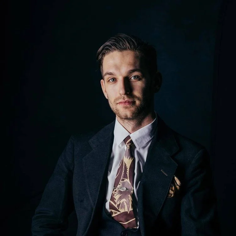 A young man with light skin and brown hair, styled with a side part, wearing a dark suit jacket, a white shirt, and a patterned tie, sitting against a dark background.