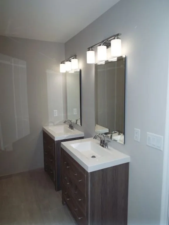 Two separate bathroom vanities. Two mirrors with  separate light fixtures over each bathroom sink. Light switches and electrical outlets are on the walls by each sink. Separate cabinets are under each vanity.
