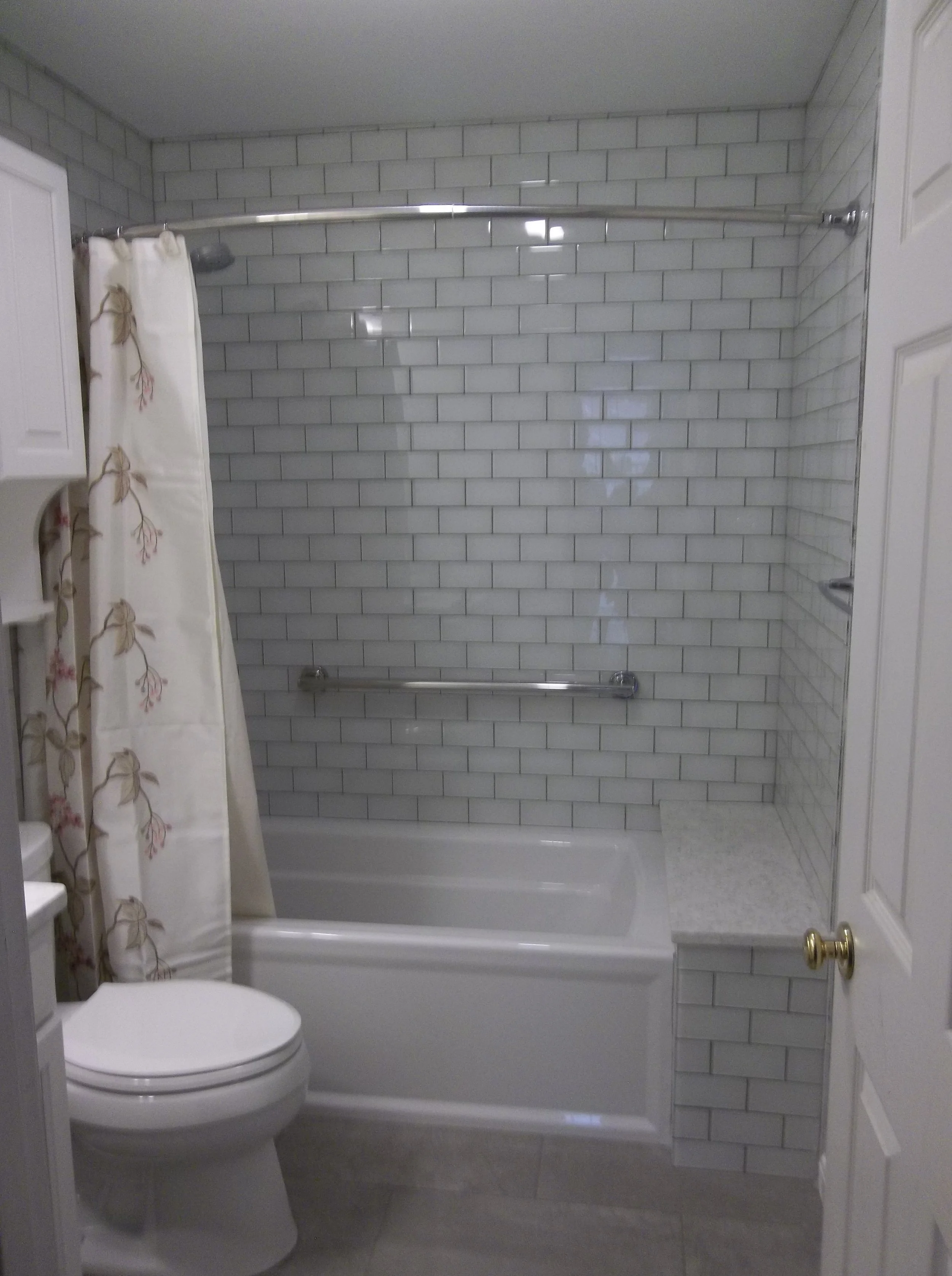Bathroom with subway tile shower area over a full sized tub. Silver shower rod with shower curtain with large flower print. White toilet on left side of photo, open door on right side of photo.