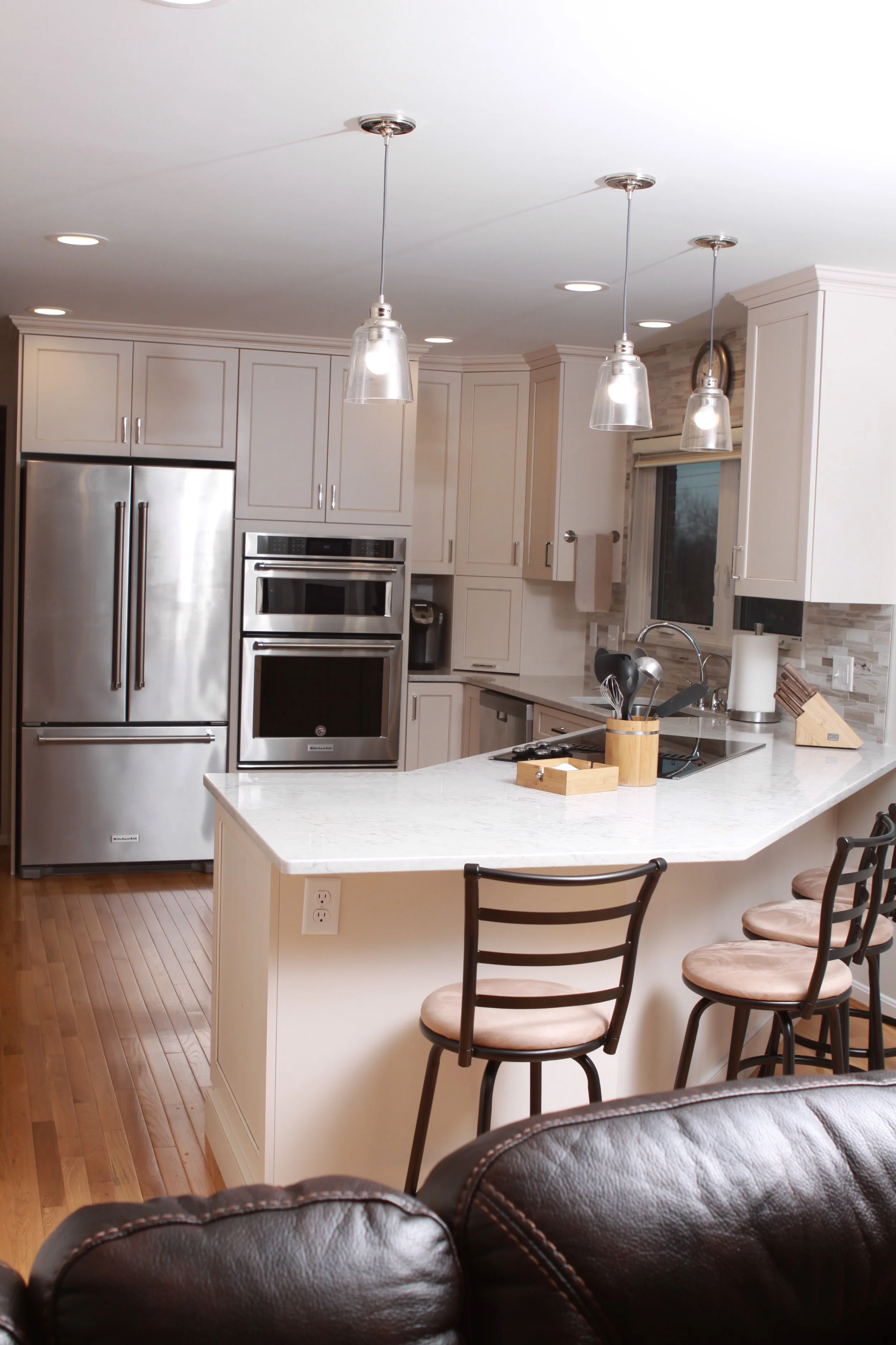 View into updated kitchen. Stainless steel refrigerator and double oven. White cabinets and  white breakfast bar with dark-backed barstools. The back of a dark leather couch in the foreground.