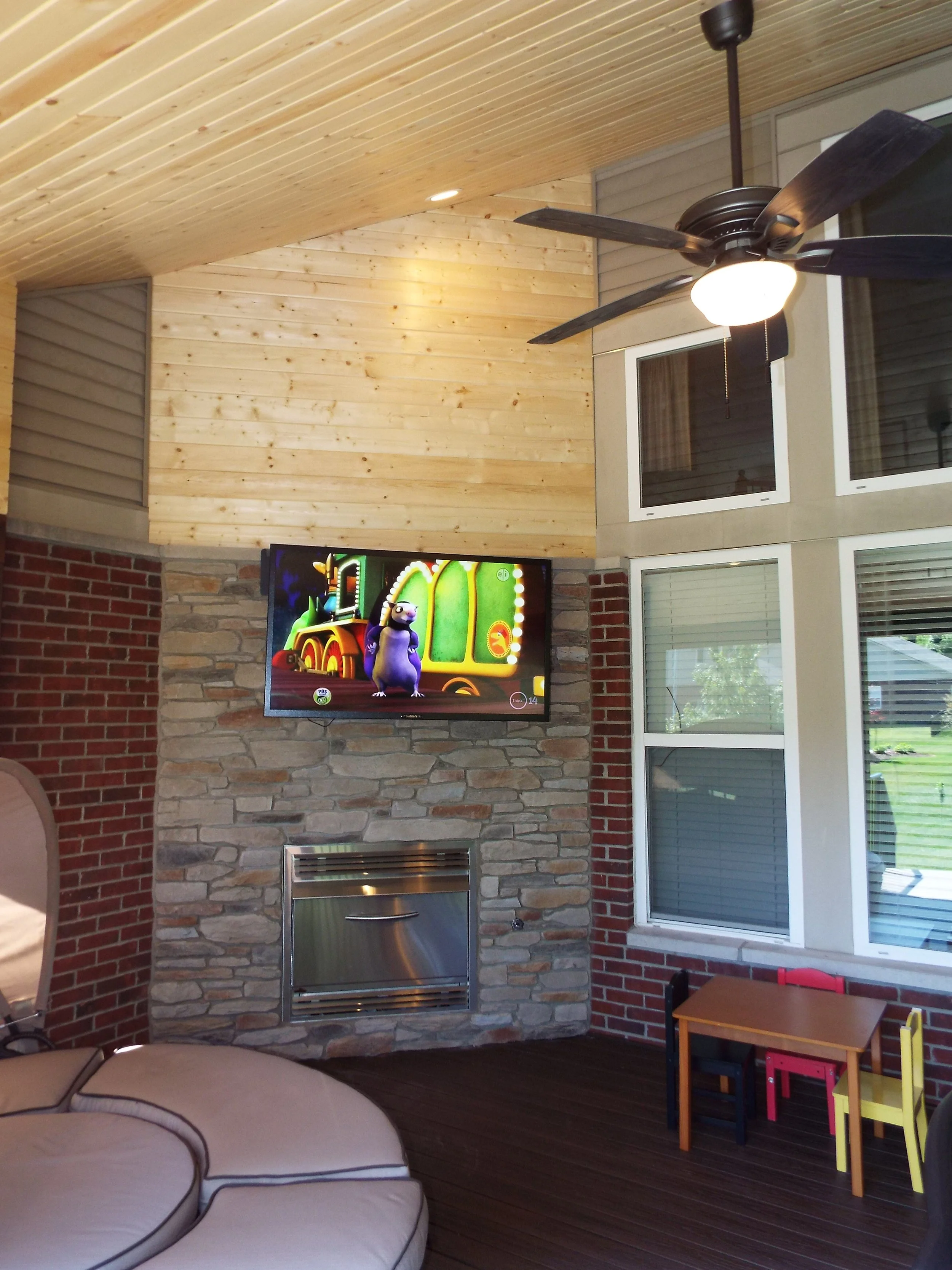 Screened in porch. Wooden bead board ceiling and upper walls. Recessed lighting and ceiling fan. Brick column and stacked stone wall.  TV mounted on stone wall. Fireplace on stone wall. Wooden floor. Round chaise lounge on left of photo. Windows from