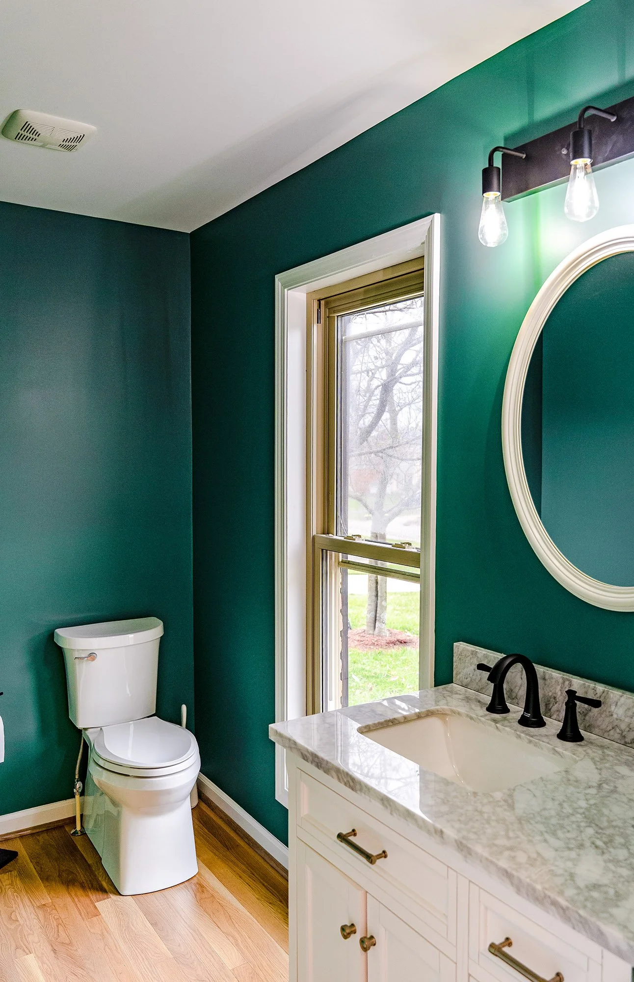 Complete First Floor Remodel by KBC Inc. 
View of bathroom area. Toilet next to large window overlooking yard. Vanity with LED light fixture and oval mirror mounted over marble sink with dark faucet and handles and light cabinets.