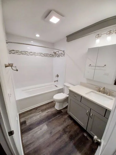 Bathroom remodeled by KBC
Simple clean bathroom. Plank flooring. Large vanity with nickel faucets and handles and white square undermount sink. Light fixtures on wall over three-sectioned mirrored medicine cabinet. Lighted exhaust fan in center of ce