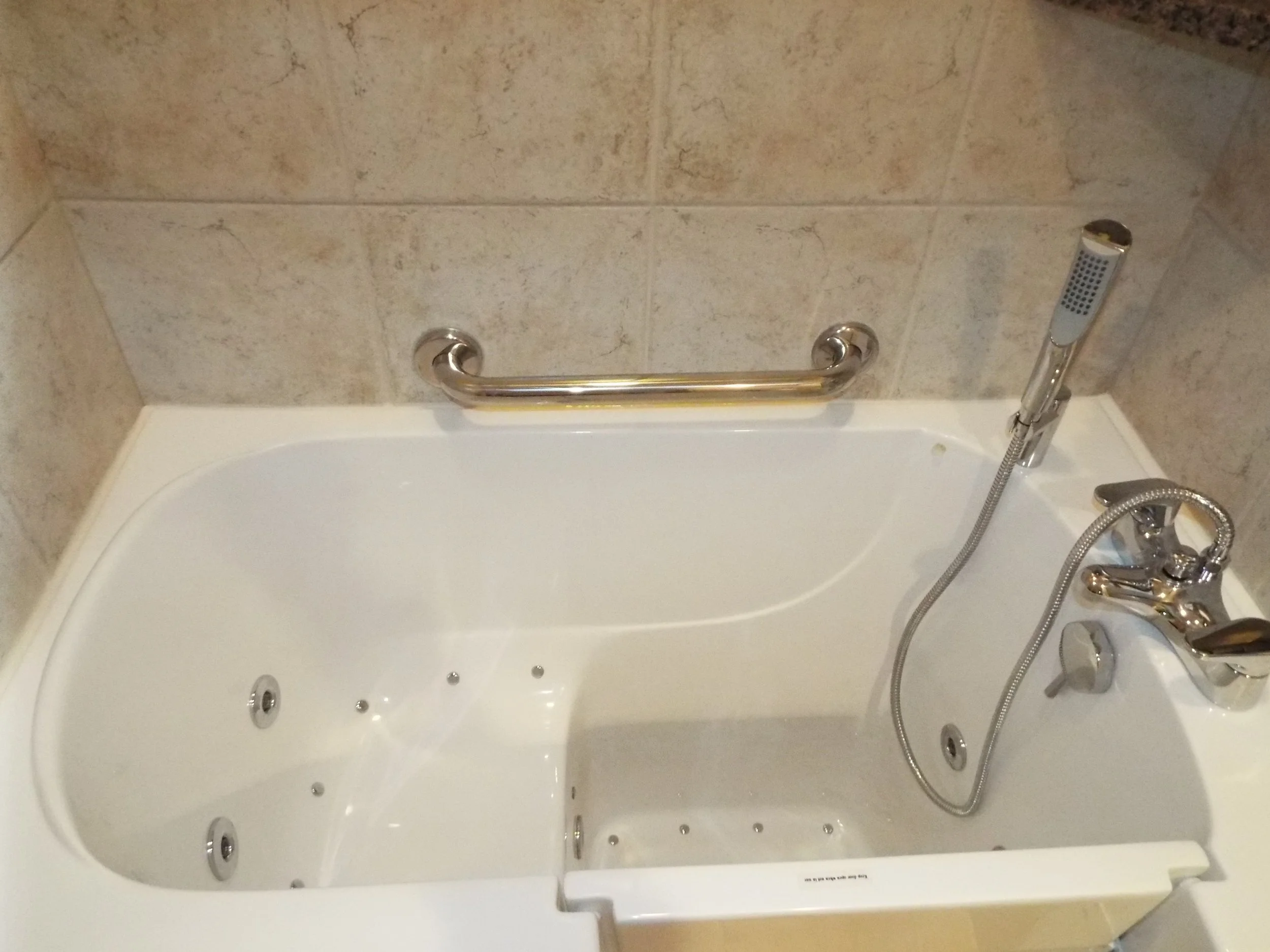 Closeup of walkin tub in a remodeled bathroom. Cararra marble on shower walls. White walk-in tub. Grab bar on shower wall. Tub faucet with connected hand-held spray. Water jets and air jets visible in tub floor and tub seat. Tub step-in door in wall 