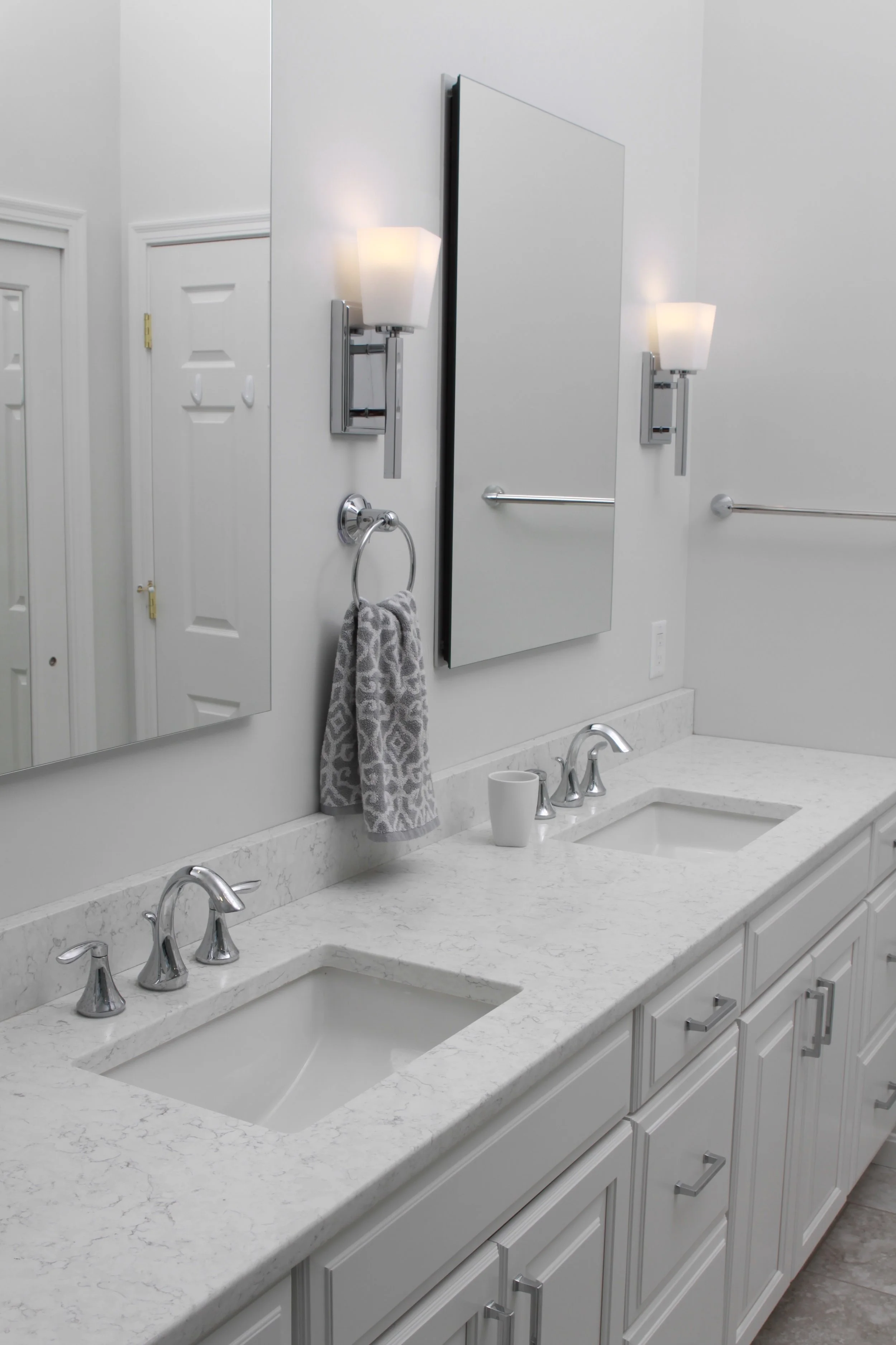 Light and airy white bathroom with double vanity sinks, silver faucets and a mirror over each sink. Two lights on the back wall, next to each mirror. Towel bar on the wall in the right side of photo.
