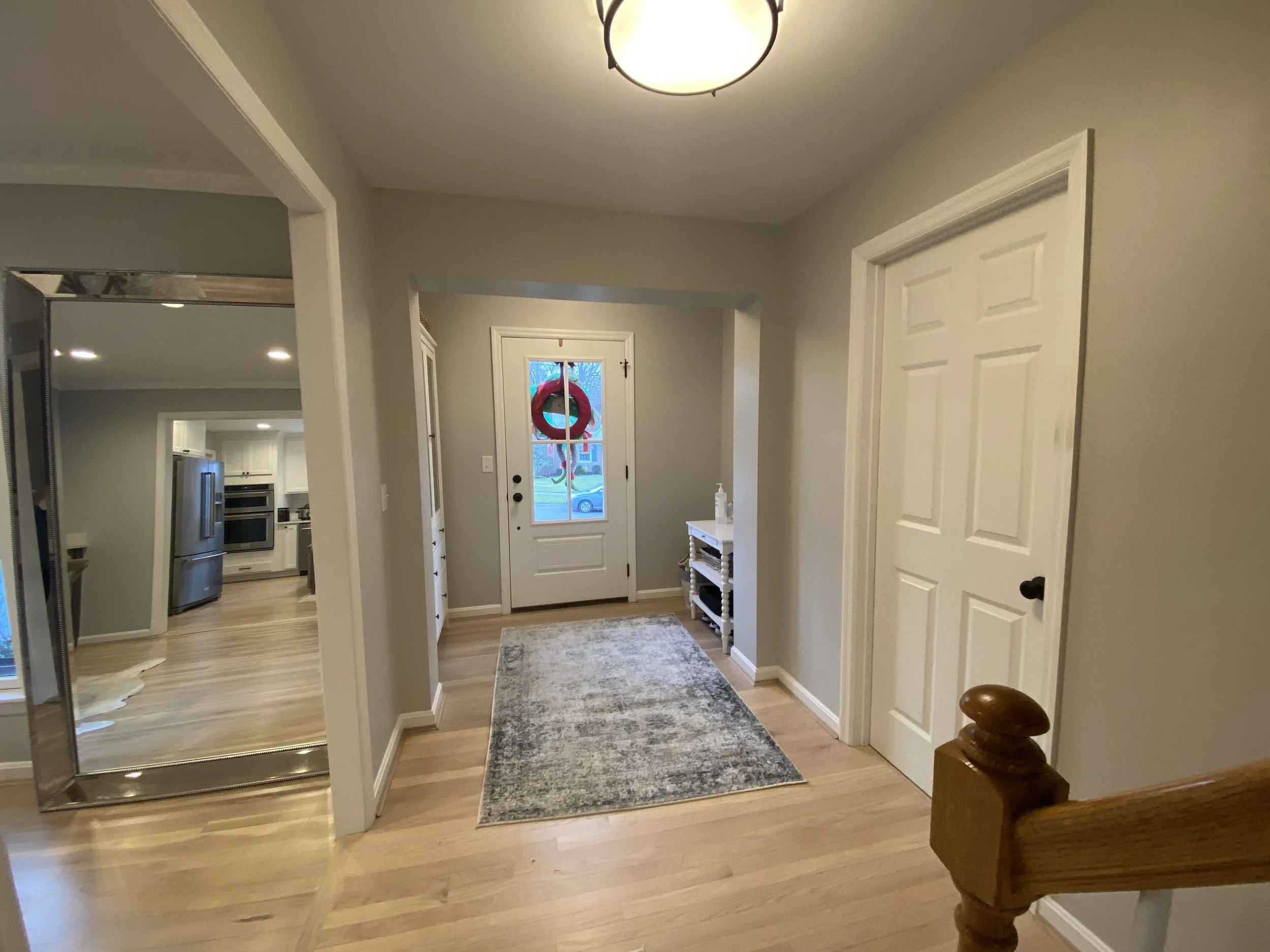 Open entryway with hardwood floor and large rug. Round lighting fixture. Stair railing on right side of photo. Large mirror showing more of the house seen through doorway on left side of photo.