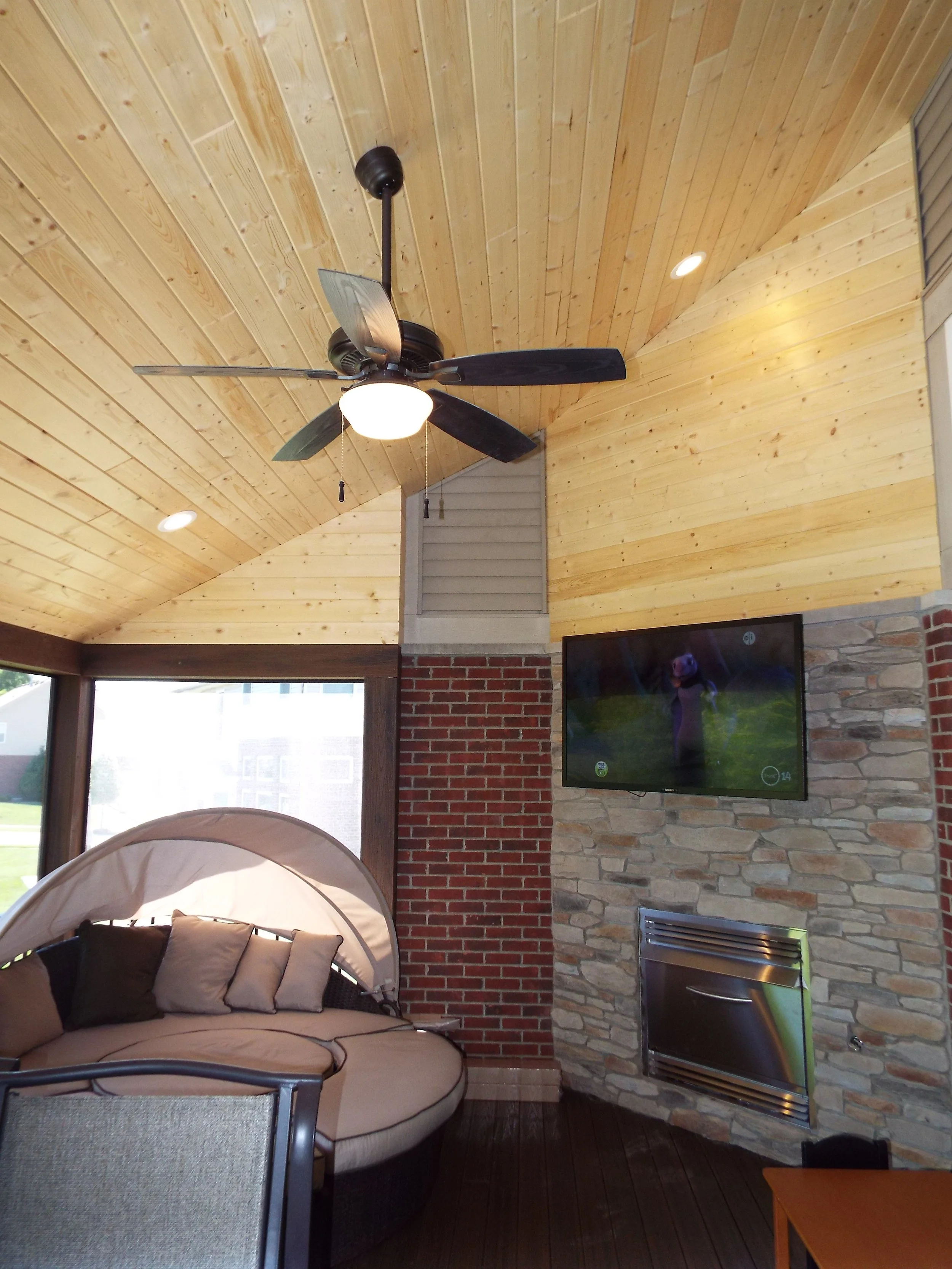 Screened in porch. Wooden bead board ceiling and upper walls. Recessed lighting and ceiling fan. Brick column and stacked stone wall. Screened windows look out onto neighboring houses. TV mounted on stone wall. Fireplace on wall. Wooden floor. Round 