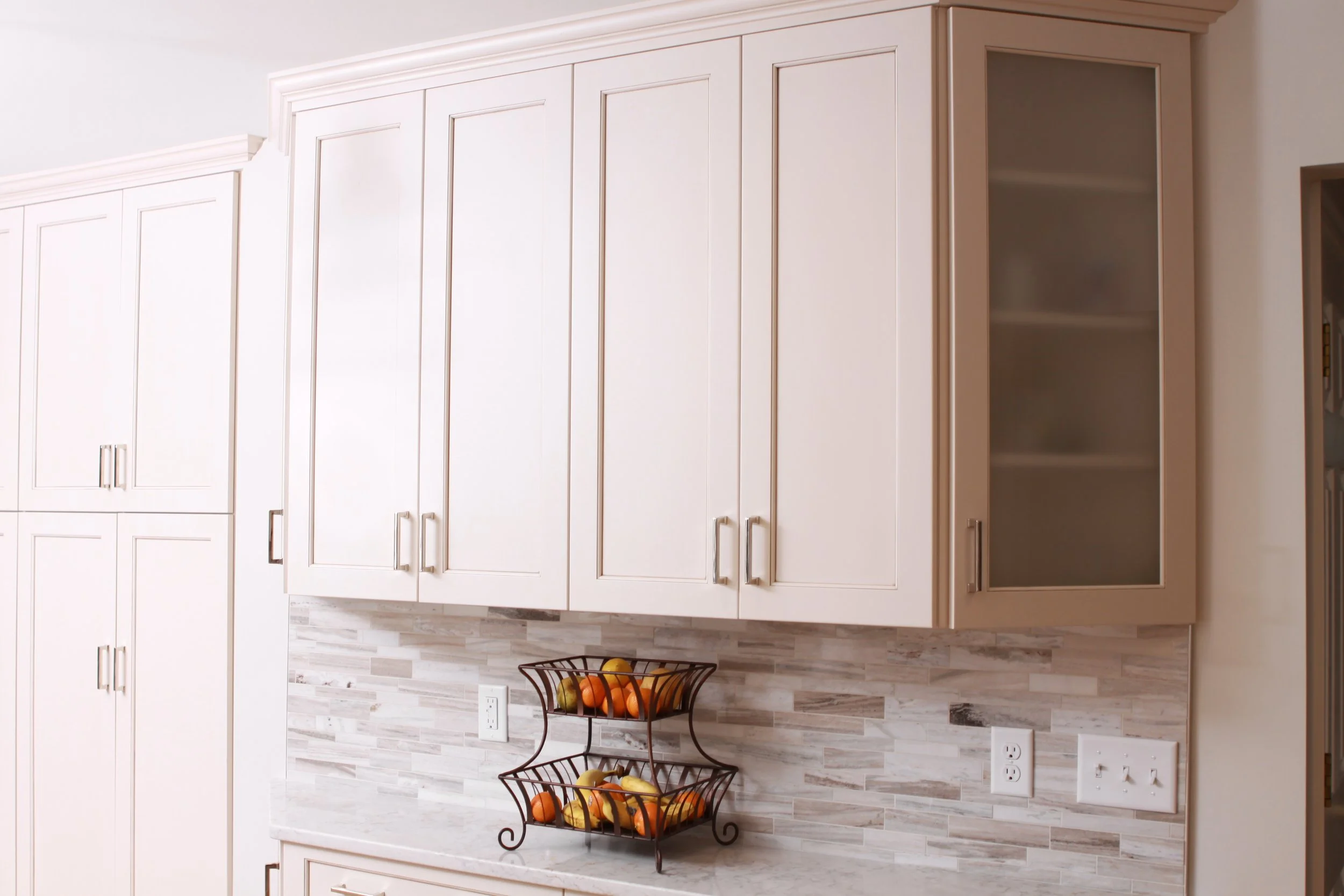 White cabinets with a grey countertop and grey tile backsplash. Basket of fruit on the countertop.