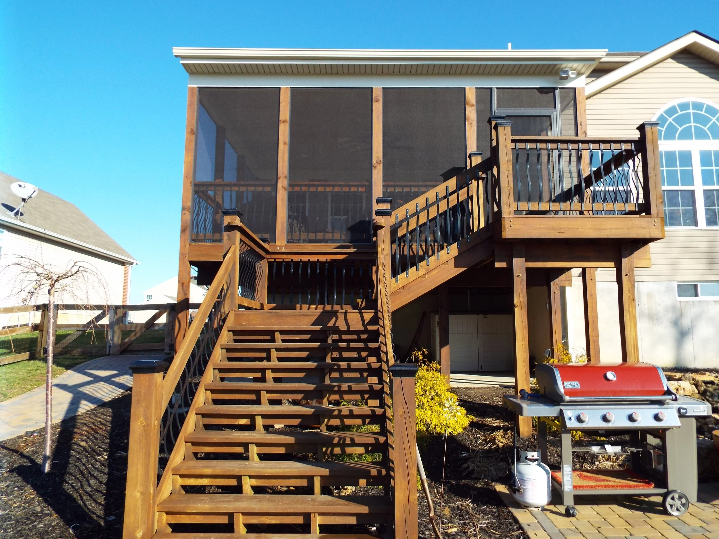 Wooden steps to screened-in porch. Large gas grill on brick patio, and dark mulch on the ground around the steps and porch posts. Screened-in porch and outside of house. Small tree and driveway on left side of photo.