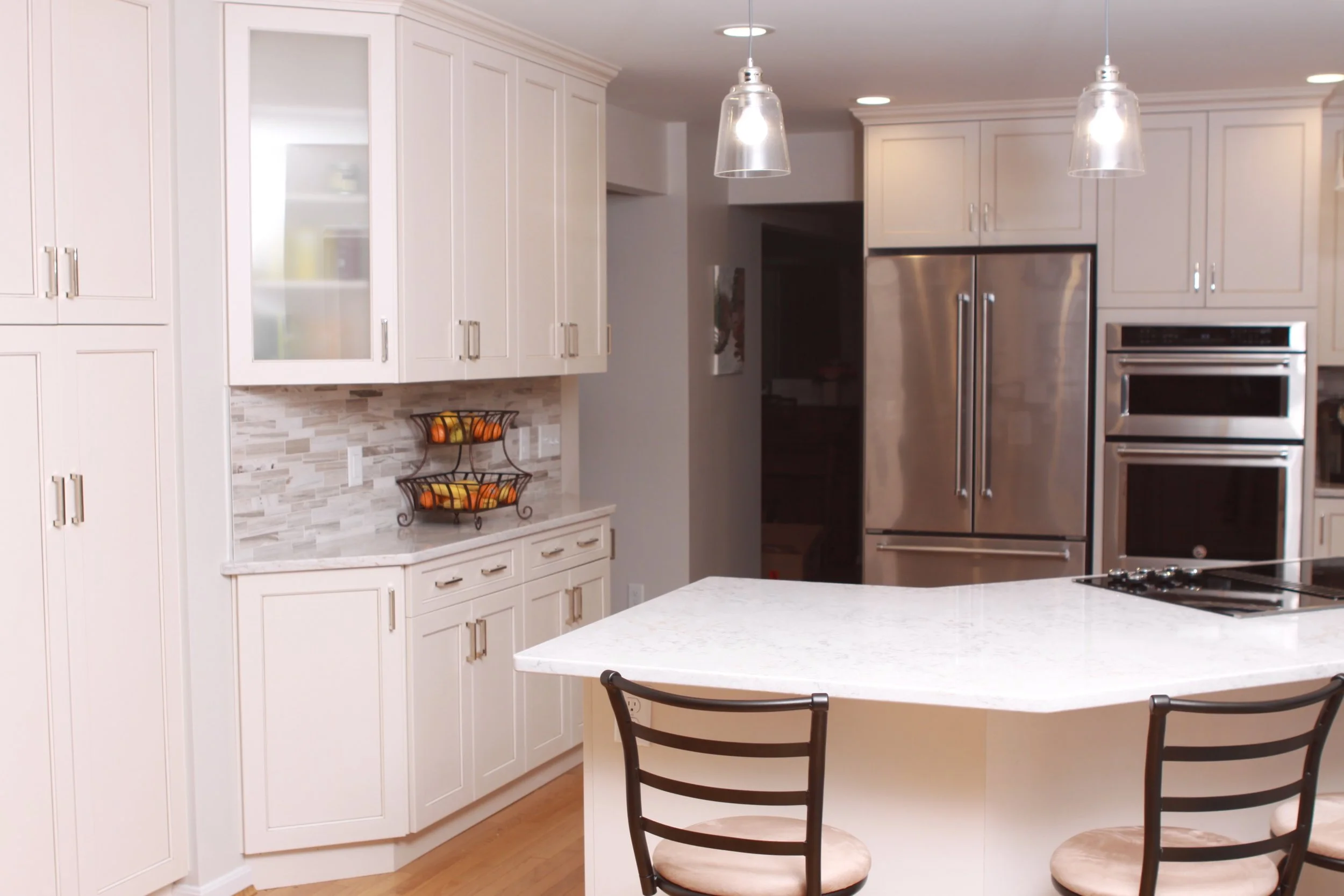 View into updated kitchen. Stainless steel refrigerator and double oven. White cabinets with a grey countertop on the left,  and  white breakfast bar with dark-backed barstools in the foreground.