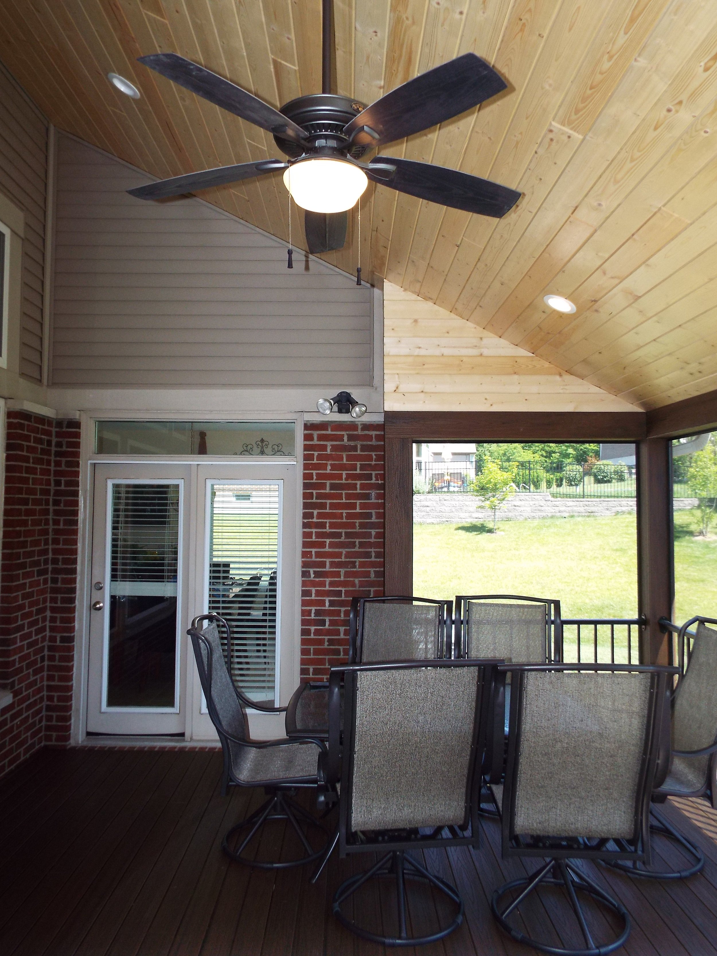 Screened in porch. Wooden bead board ceiling. External house siding and brick walls from exterior of house. Recessed lighting and ceiling fan. TV mounted on stone wall. Fireplace on stone wall. Wooden floor. Outdoor patio table and six chairs on righ