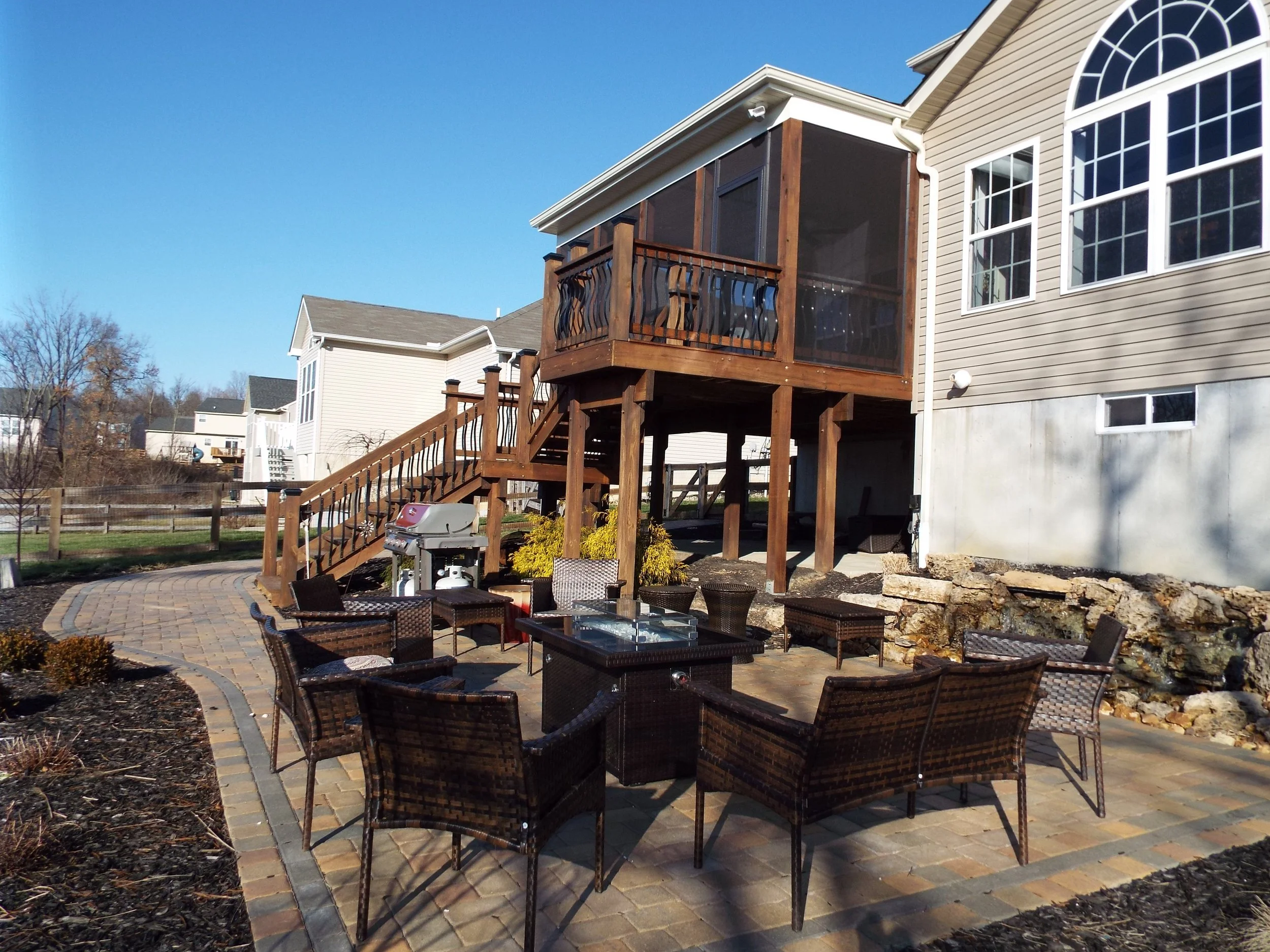 Large brick patio with dark wicker seating around firepit. Large gas grill at the base of the steps leading to the large screened-in porch. Neighboring houses in the background.
