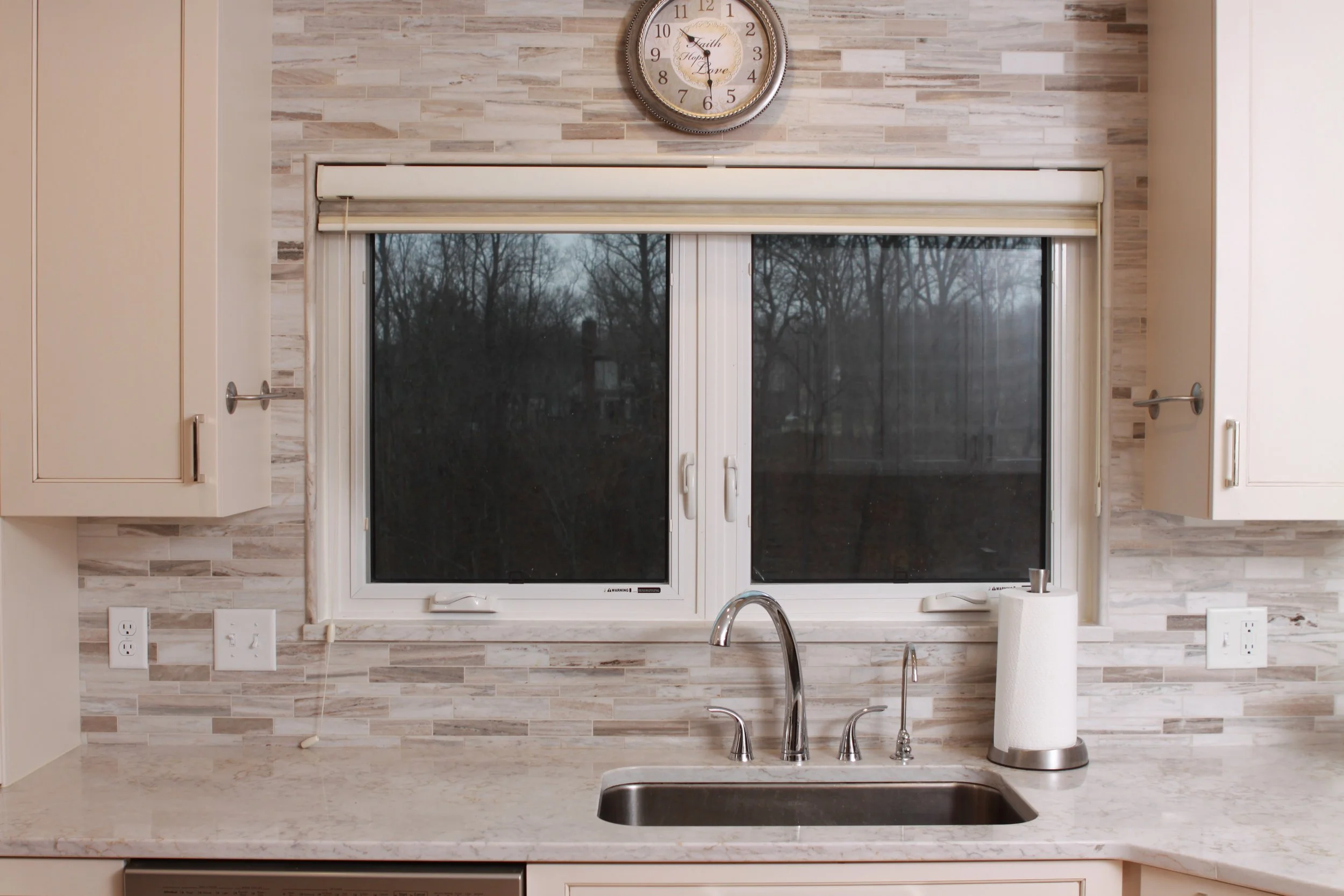 Clock over the kitchen windows with a grey tile backsplash. White cabinets on each side, light grey counter top and stainless steel sink.