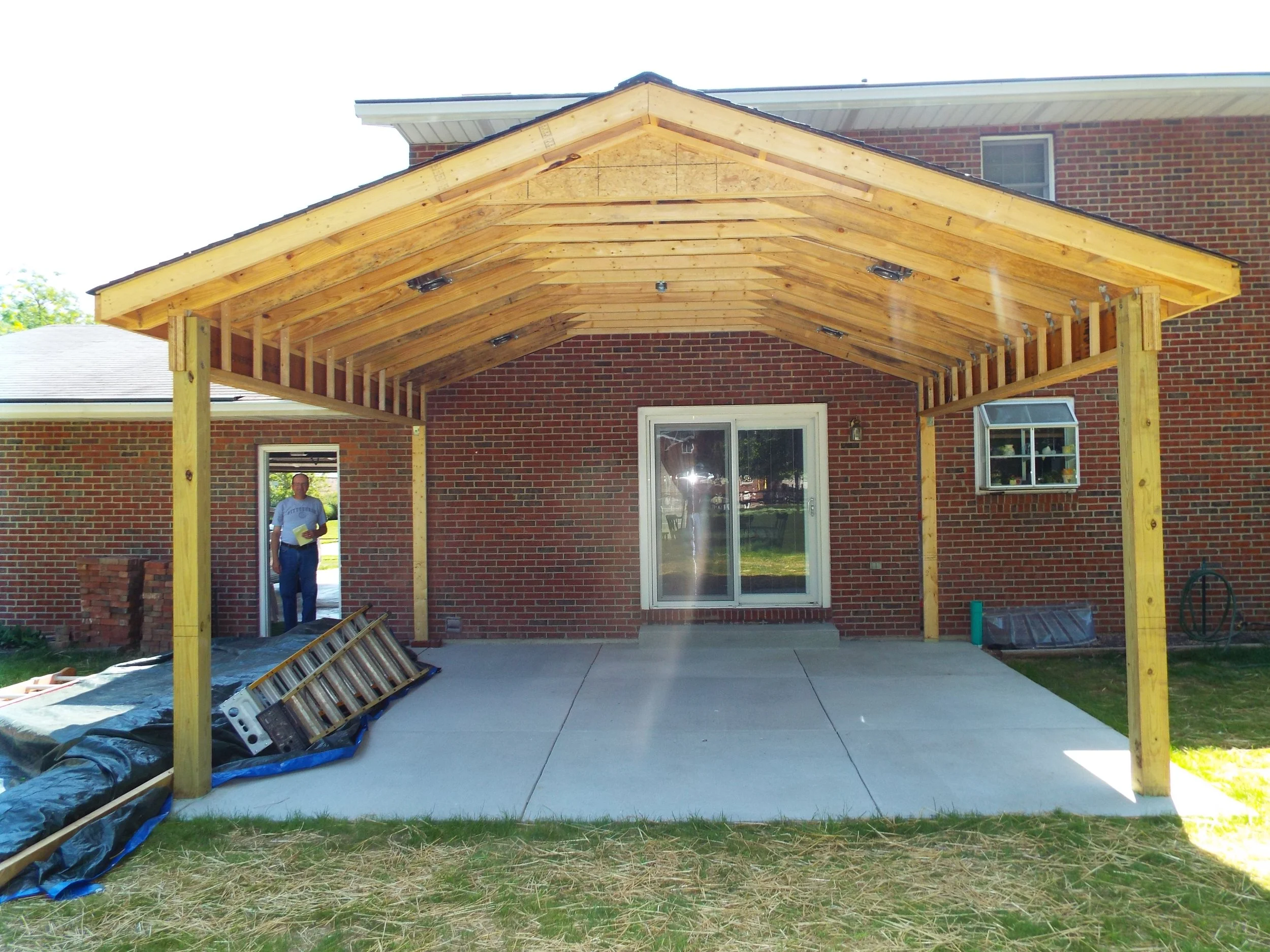 Construction on covered patio. Shingled roof over timber rafters. Two ladders sit in shade on concrete. Large tarp on left side of frame. Person standing in open doorway on left of photo. Sliding patio door reflects yard in center of photo.