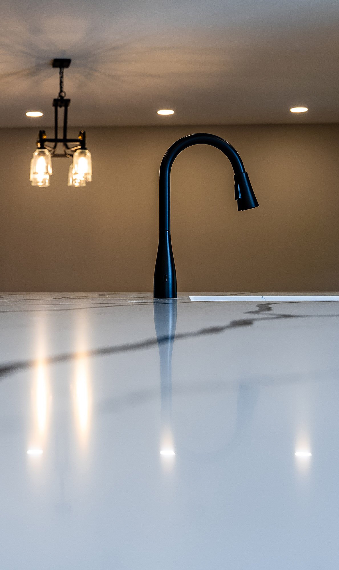 Complete First Floor Remodel by KBC Inc. Closeup view of sink faucet, mounted on marble kitchen island. LED can lights mounted in ceiling, and hanging chandelier on the left side of photo.