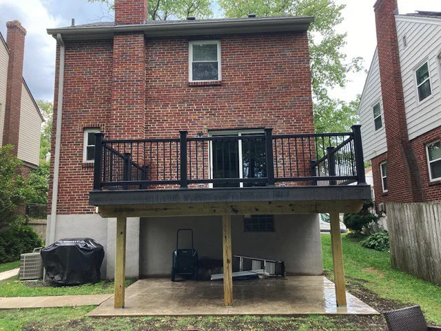 KBC Inc.  New Deck and Patio Project. This photo shows a Large two-story brick house with  a large deck addition. The deck has dark metal railings and is sitting on wooden posts. A double sliding glass door leads into the house. The deck is covering