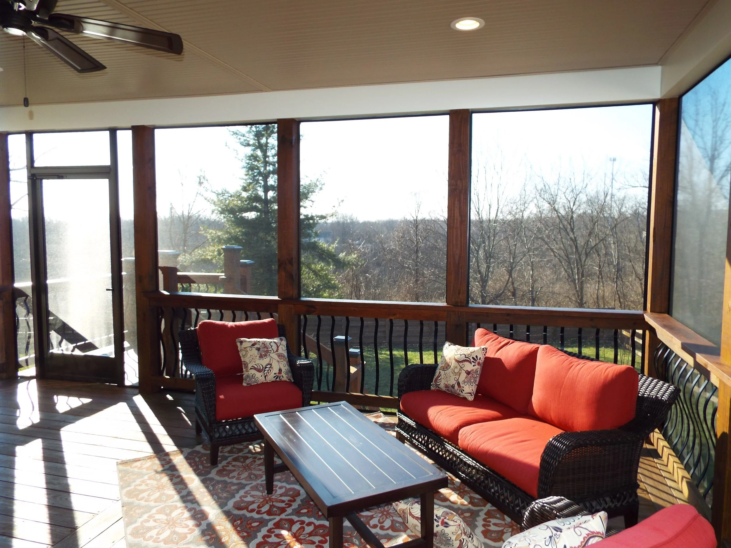 Screened-in porch overlooking trees and woods. Wicker love seat and chairs with brightly colored cushions and pillows. Wooden railing with metal posts. Brightly patterned rug over wood plank floor.