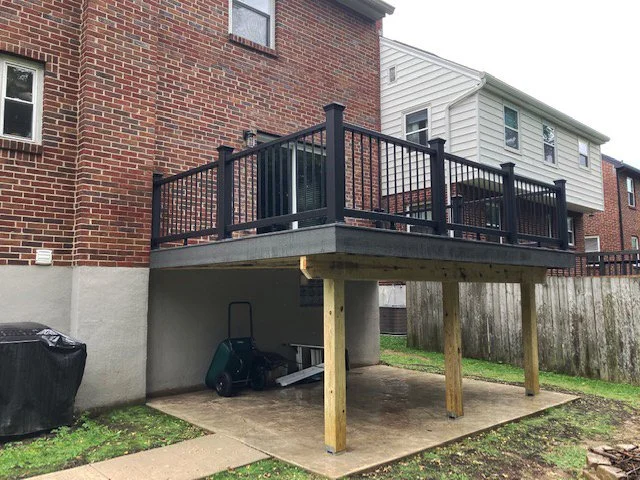 KBC Inc.  New Deck and Patio Project. This photo shows a Large two-story brick house with  a large deck addition. The deck has dark metal railings and is sitting on wooden posts. The deck is covering for a patio space. On the left side of the patio a