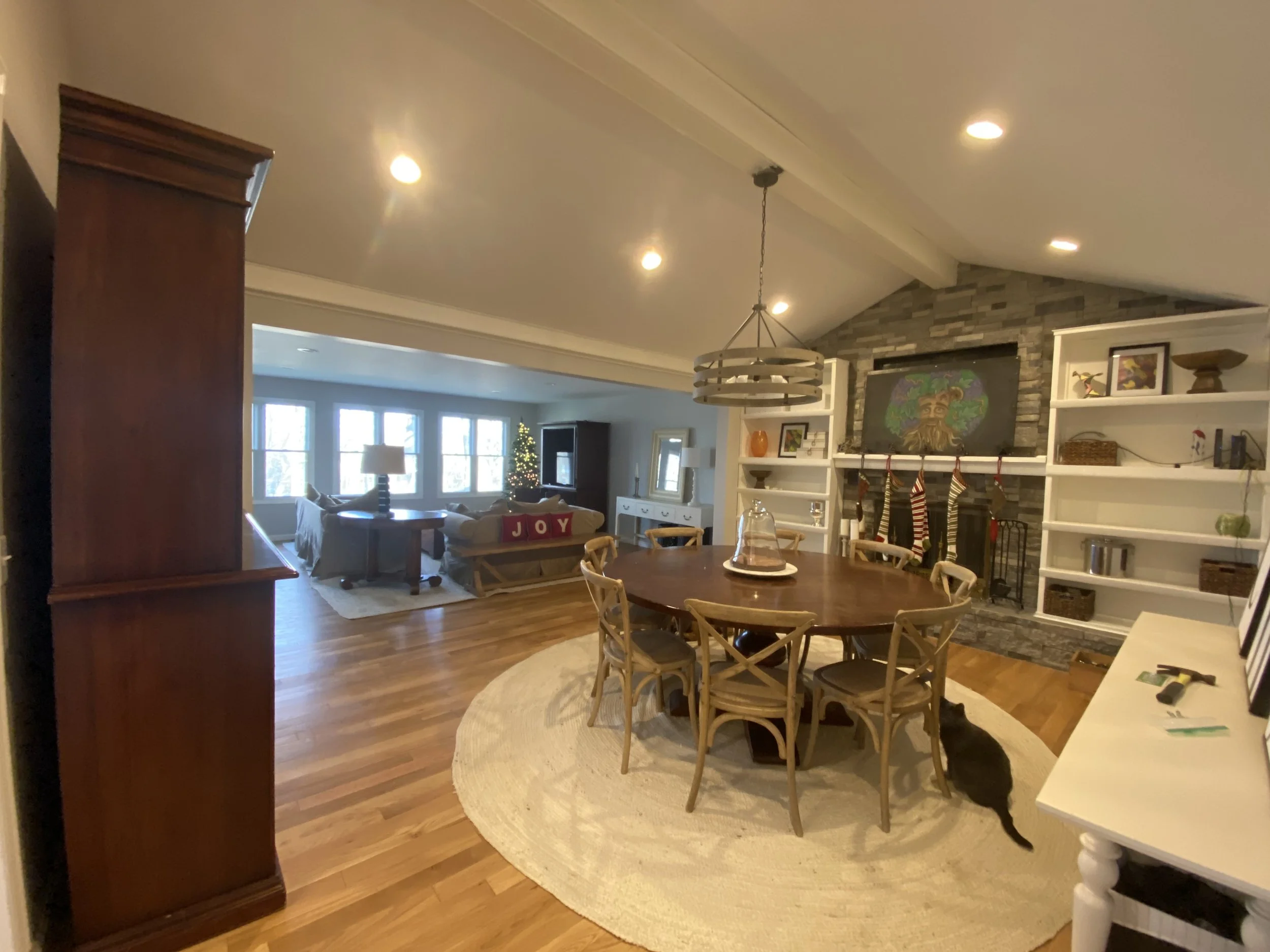 Large open-concept family room with cathedral ceilings. Recessed can lighting, and a circular chandelier over a round table with six chairs. Large round woven rug on hardwood plank floors. Bookcases on left and right of a large brick fireplace with C
