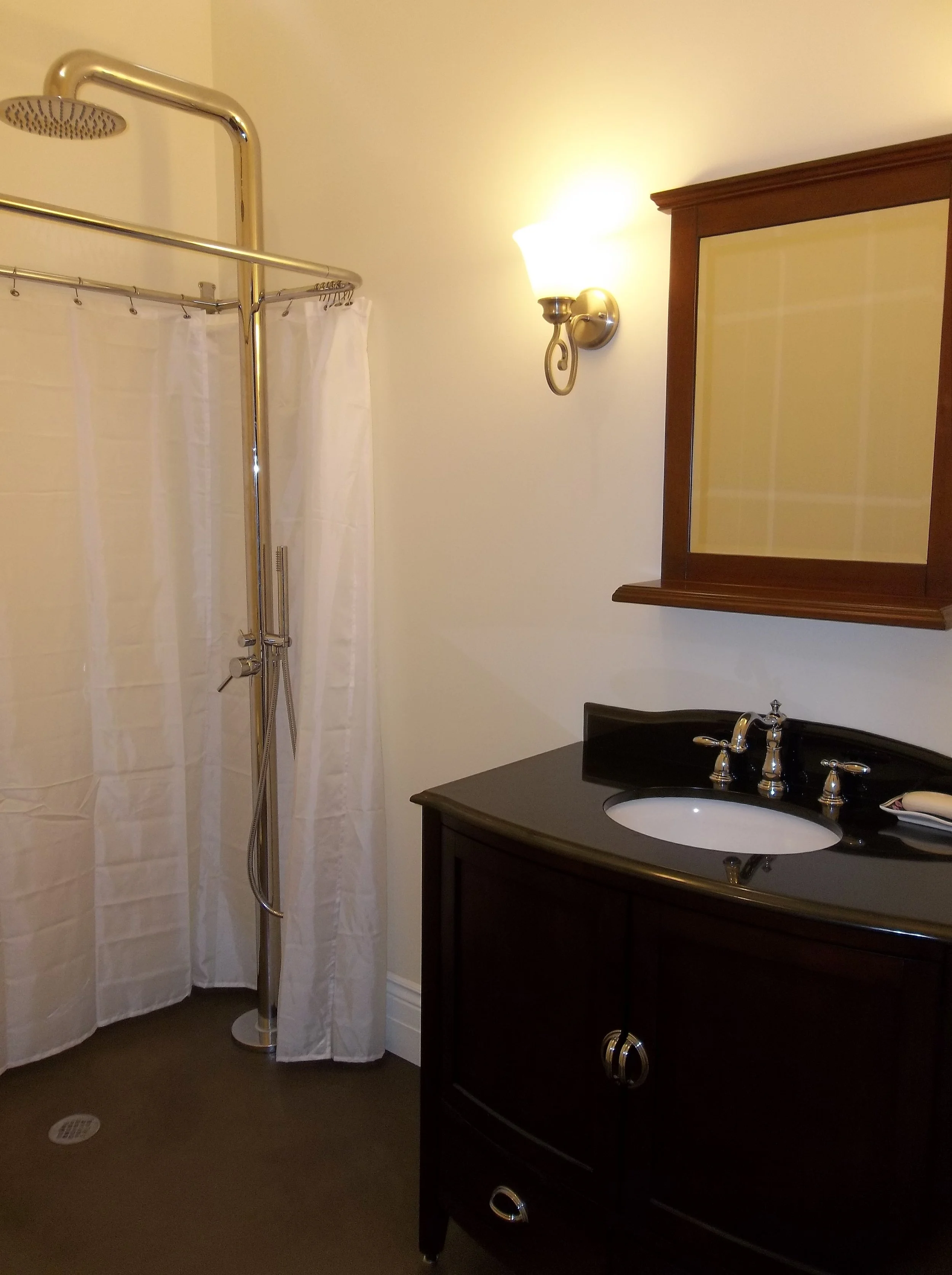 Barrier free corner shower and vanity.  Brass shower pole, shower head and curtain rod. White shower curtain partially pulled open. Stained concrete floor. Dark vanity cabinets and sinktop with white undermounted sink. Brushed nickel faucets. Dark me