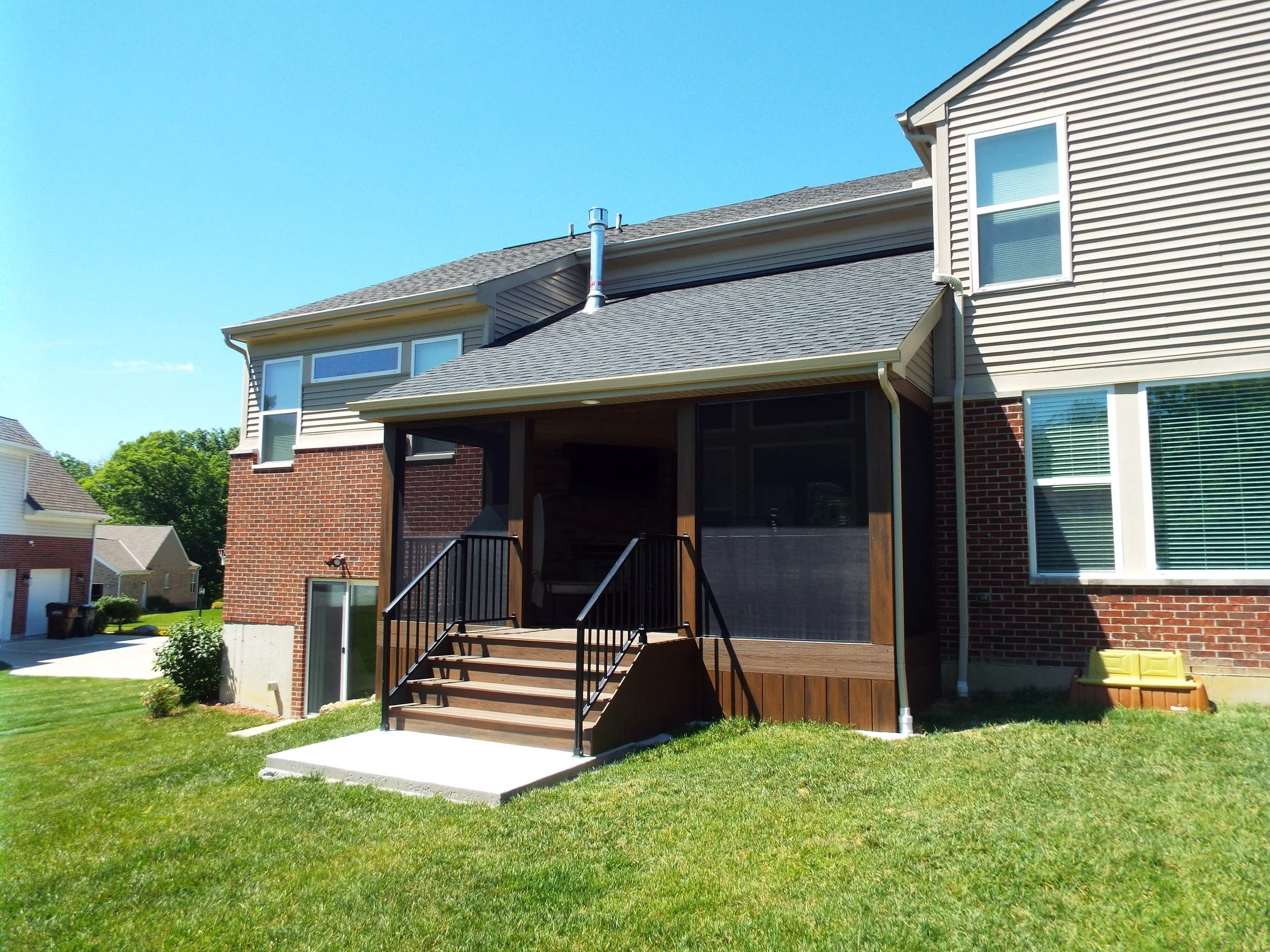 Exterior view of screened-in porch. Shingled roof. Dark stained wooden posts and stairway.  Dark stained panels covering under deck. Dark metal railing lead to square concrete pad in center of lawn. Seating on deck visible through screened windows. E