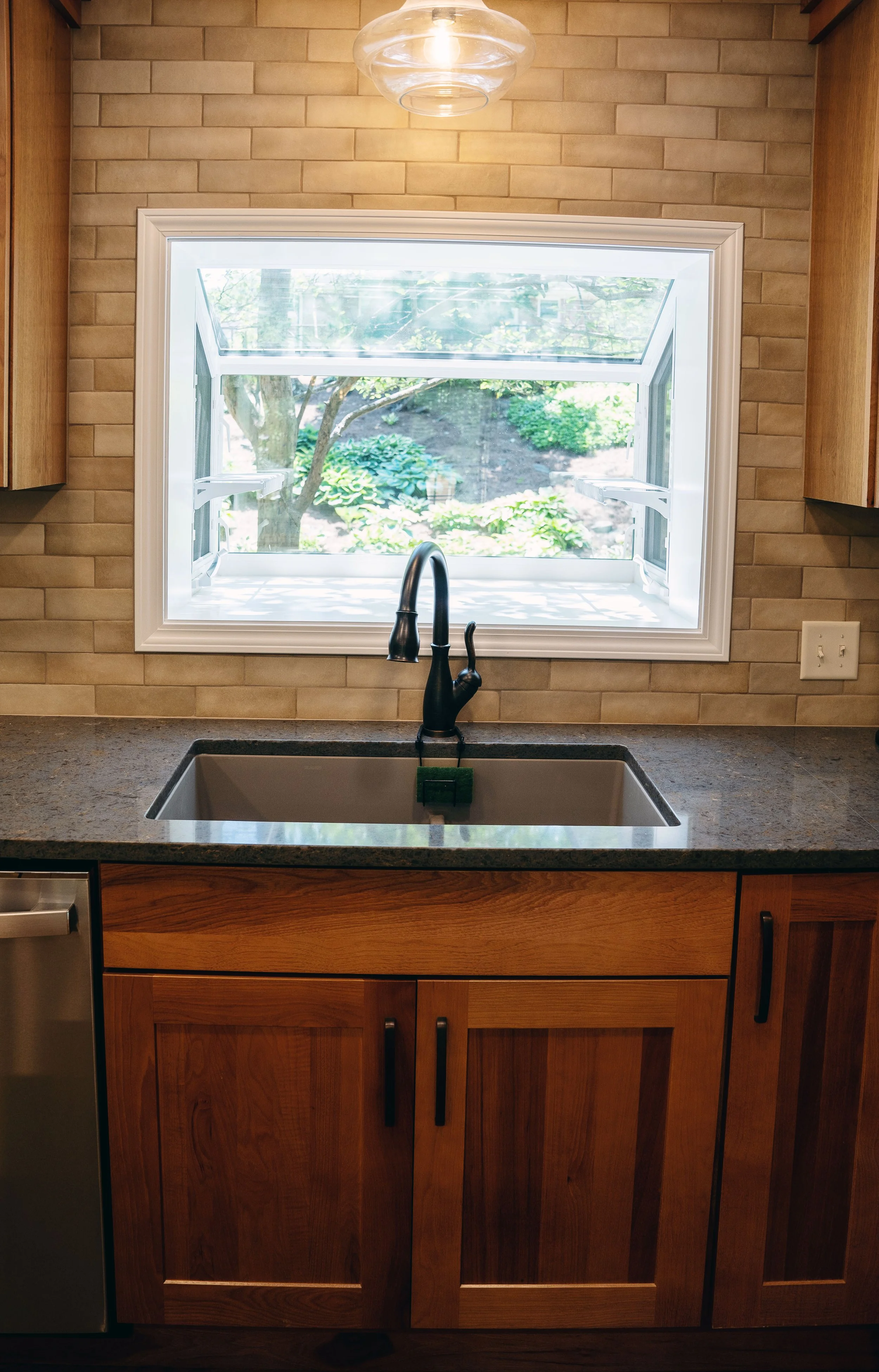 KBC Construction - Whole House Remodel in Lakeside Park, KY. Closeup of undermounted facuet with grey countertops. Dark wood cabinets. Earthtone brick blacksplash. A window looks overlooks the back yard.