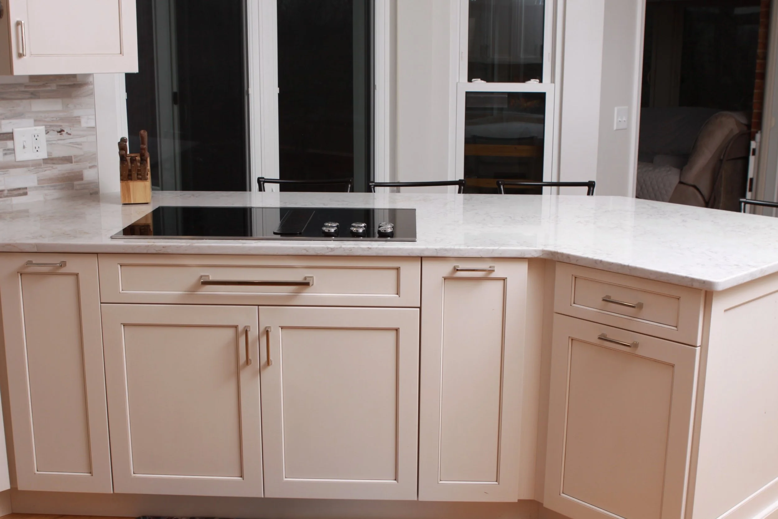 Dark electric stovetop in the center of the counter, with windows across the wall. White cabinets with a grey countertop and grey tile backsplash.
