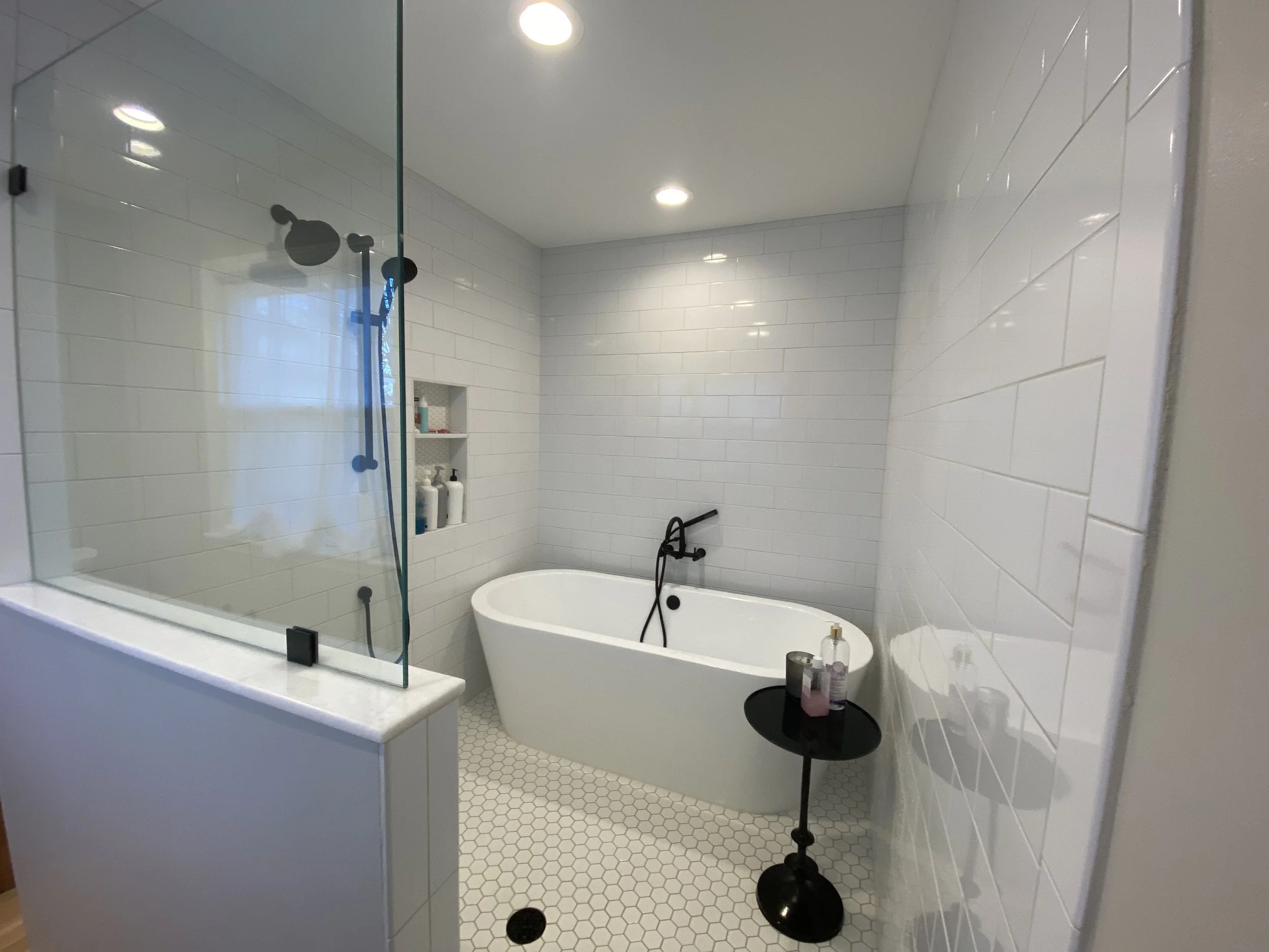 Barrier-free shower with soaking tub. Subway tile on walls and hexagonal tiles on floor. Recessed can lighting on ceiling. Solid glass half-wall. Black fixtures. Dark round table holding soaps and a candle on right side of tub.