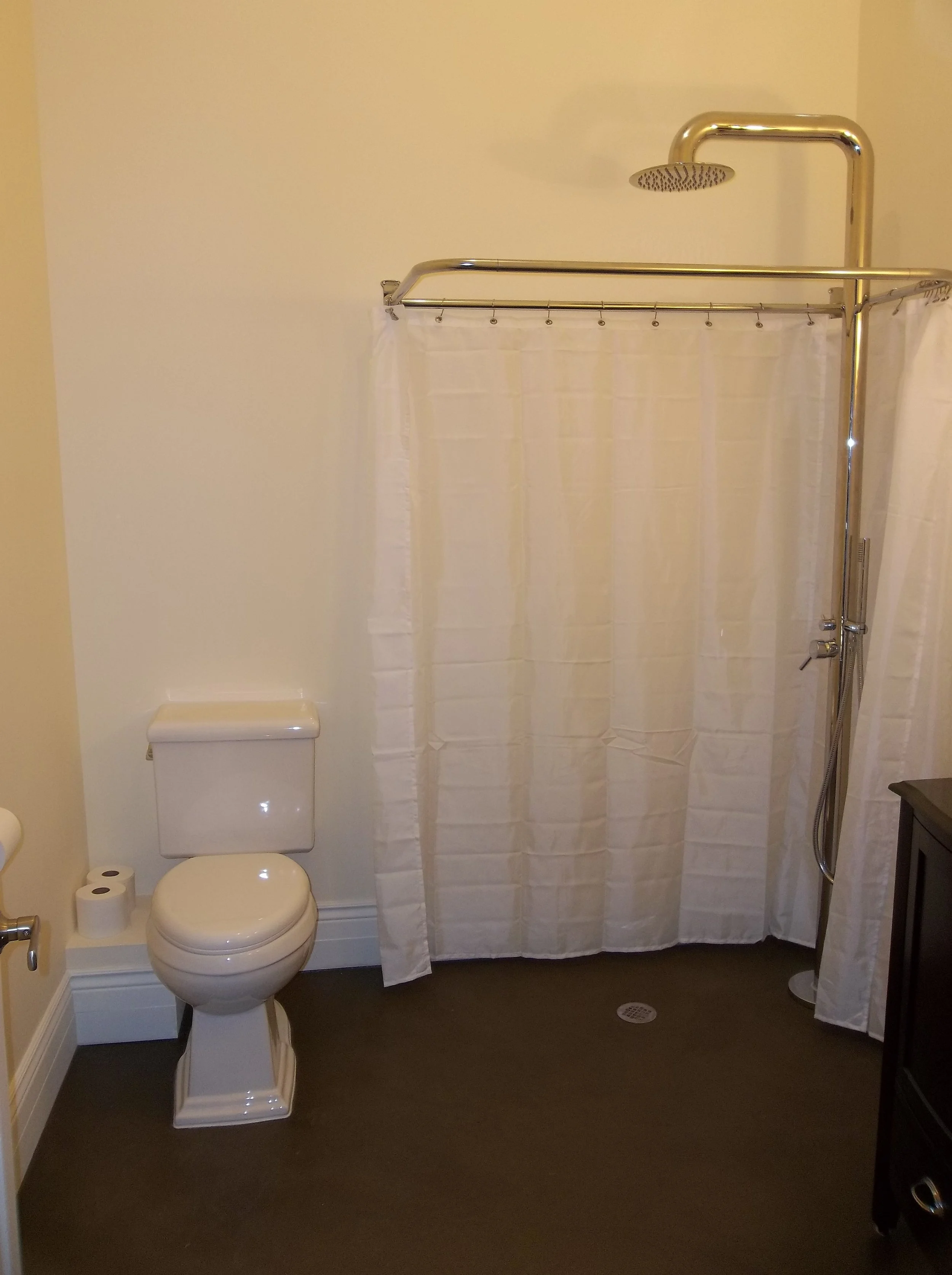 Barrier free bathroom.  Brass shower pole, shower head and curtain rod. White shower curtain partially pulled open. Stained concrete floor. White toilet on left side of bathroom.
