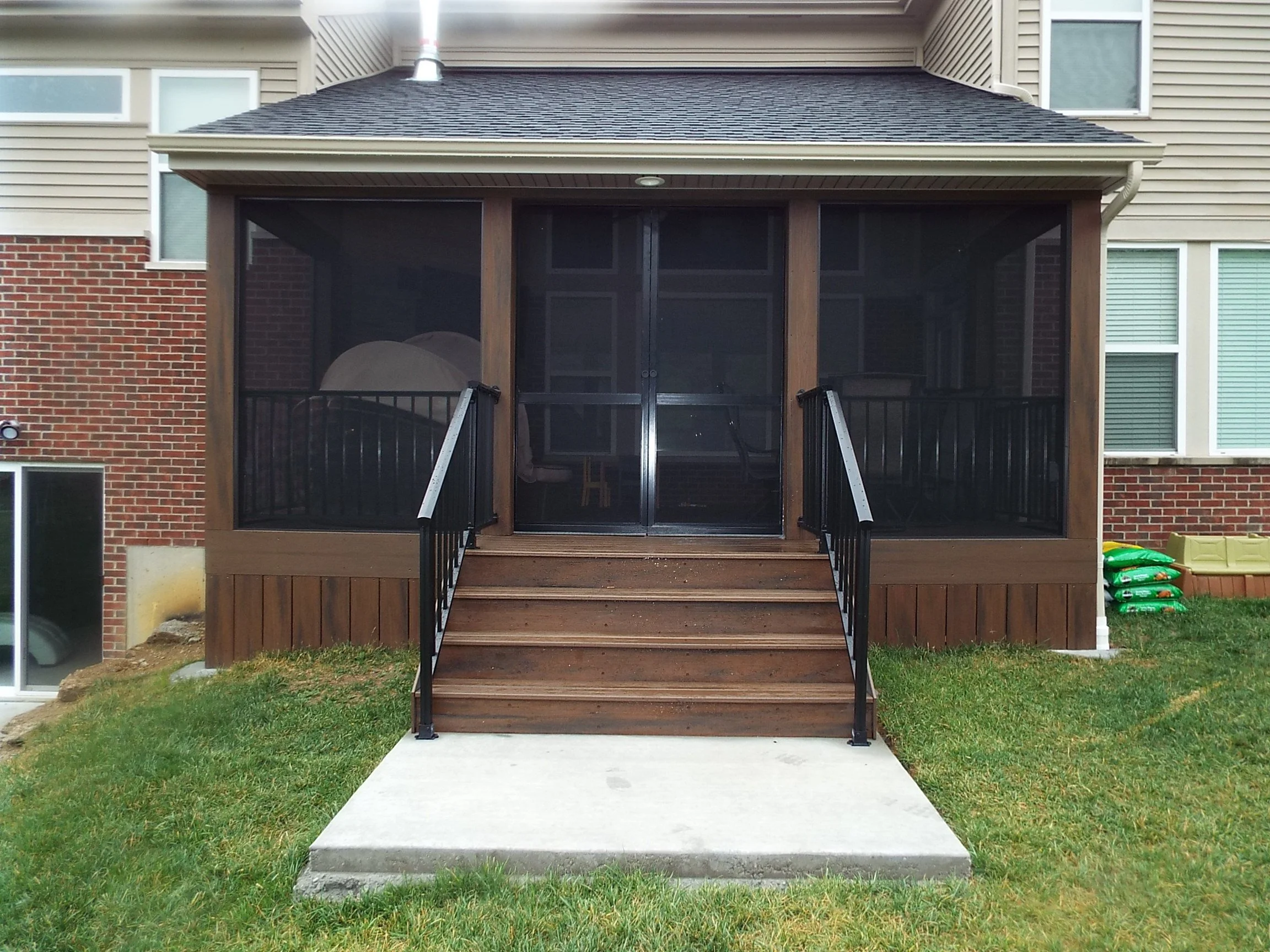 Exterior view of screened-in porch. Shingled roof. Dark stained wooden posts and stairway.  Dark stained panels covering under deck. Dark metal railing lead to square concrete pad in center of lawn. Seating on deck visible through screened windows.