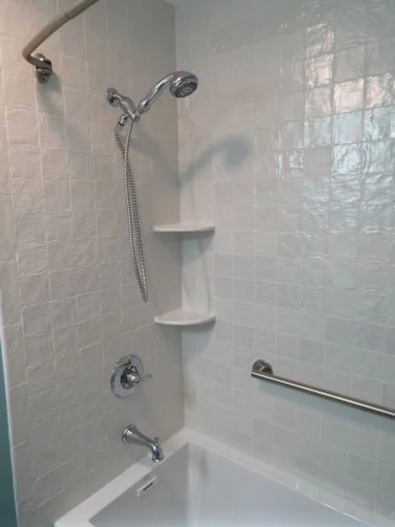 Close up on interior of shower. Corner shelves next to the hand-held shower sprayer. There is a grab bar on the back wall of the shower.