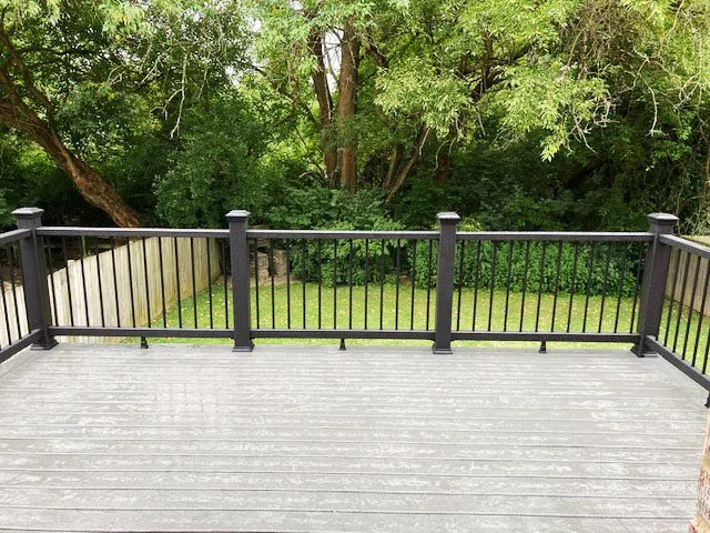 KBC Inc.  New Deck and Patio Project. This photo shows a composite deck with dark metal railings. The deck overlooks a back yard. A wooden fence is visible on the left side of the yard, and bushes and trees are growing across the back of the yard.