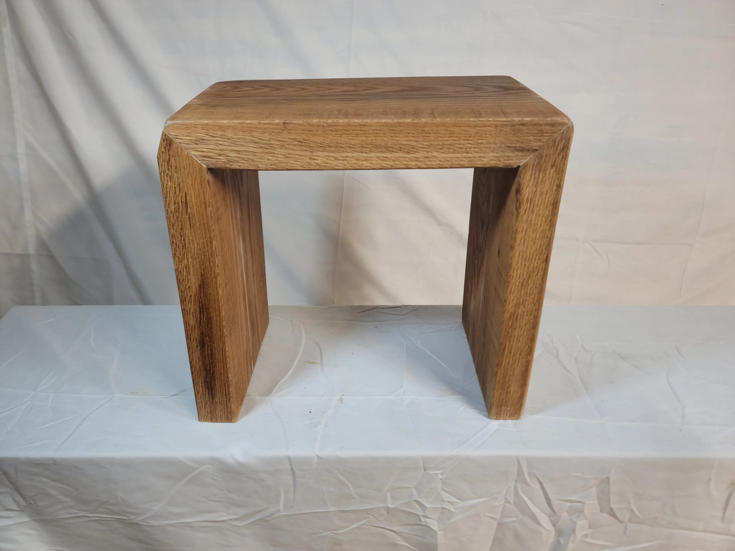A wooden stool with a curved top and two legs, placed on a white surface with a plain background.